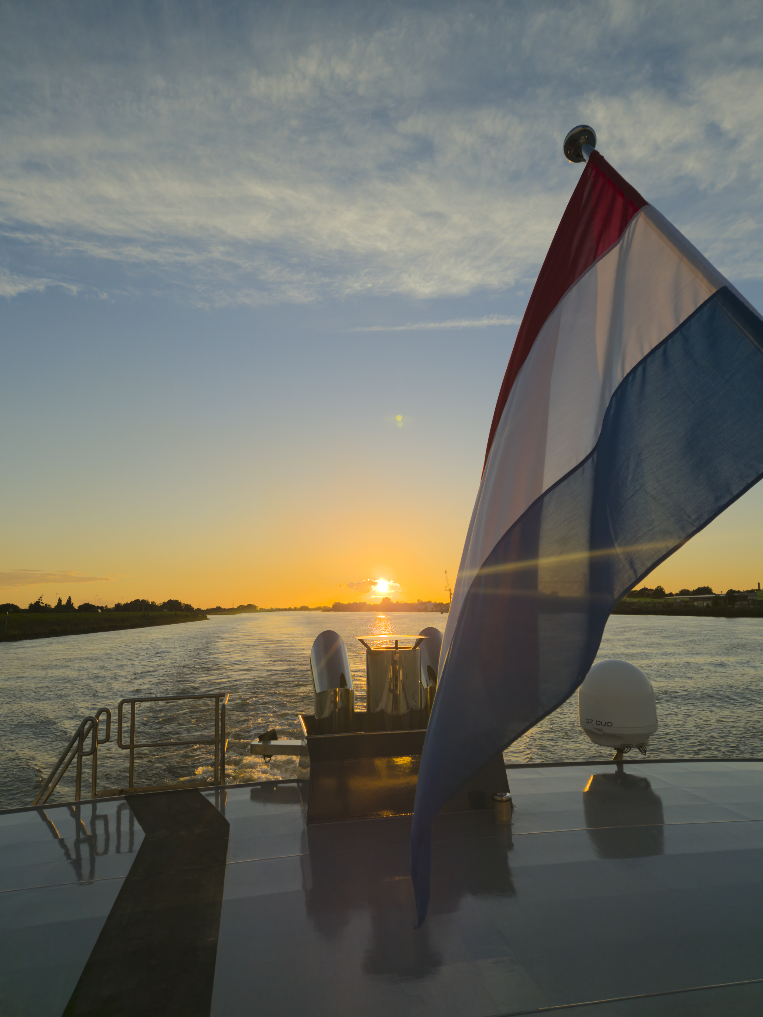 Een vlag met de kleuren rood, wit en blauw wappert in de wind op een boot tijdens zonsondergang, met de rivier en de ondergaande zon op de achtergrond.