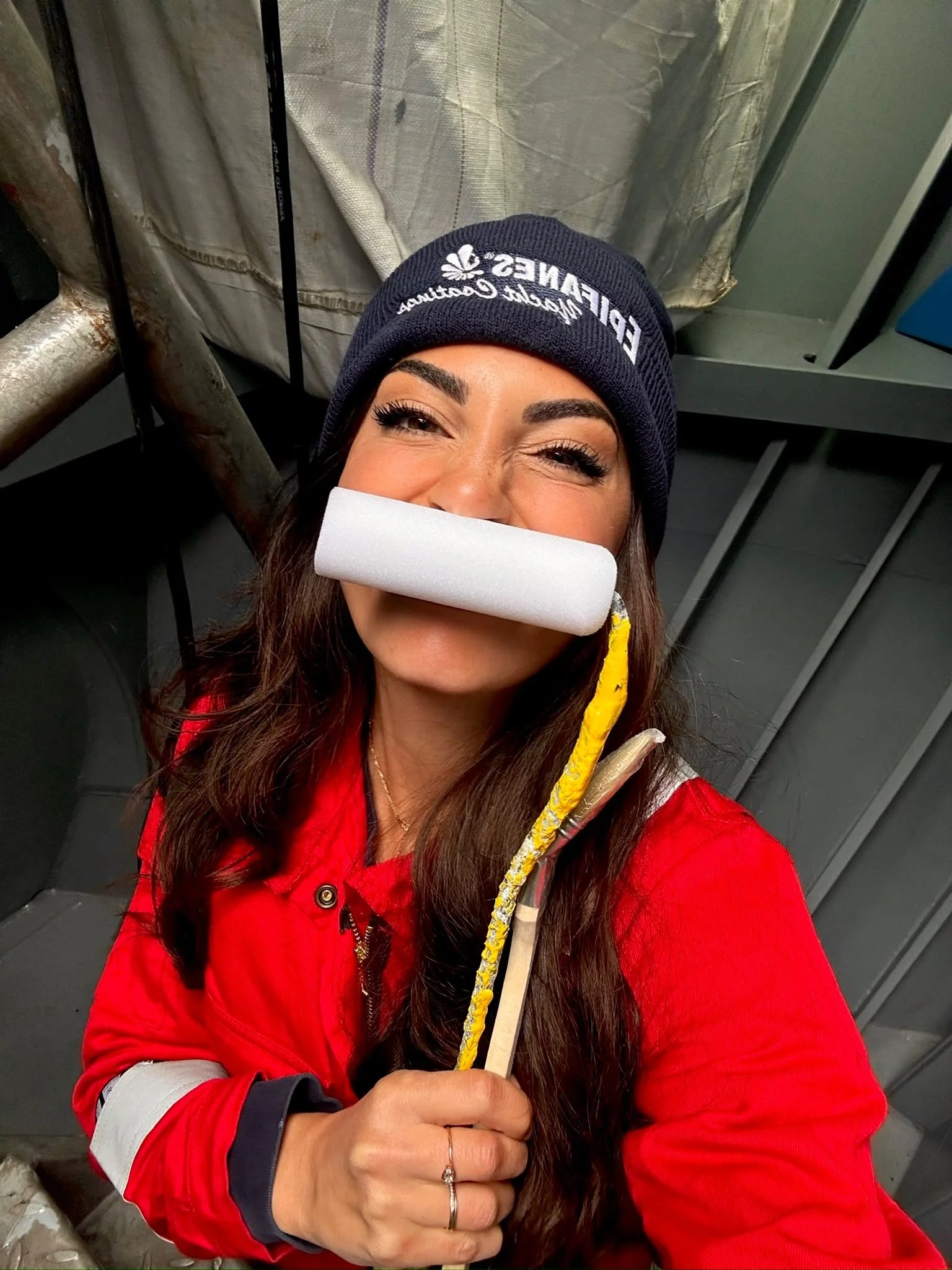 Een vrouw met donker haar en wenkbrauwen draagt een blauwe muts met witte tekst en een rode jas. Ze houdt een geverfde stok vast en knipoogt naar de camera. Op de achtergrond is een metalen structuur.