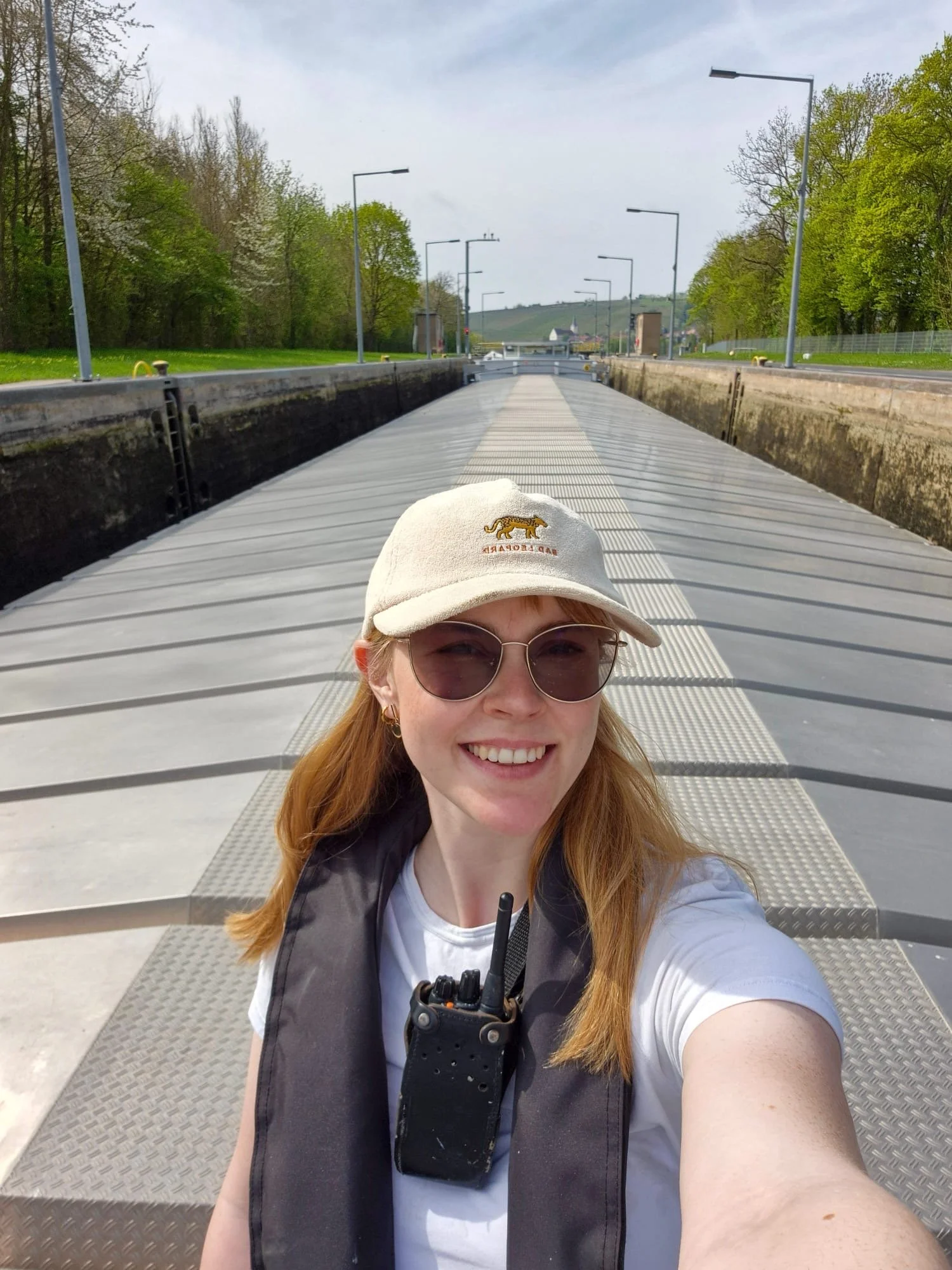 Vrouw met rode haar, zonnebril, een baseballpet met een leeuwlogo en een backpack, neemt een selfie op een brug met een berglandschap op de achtergrond.