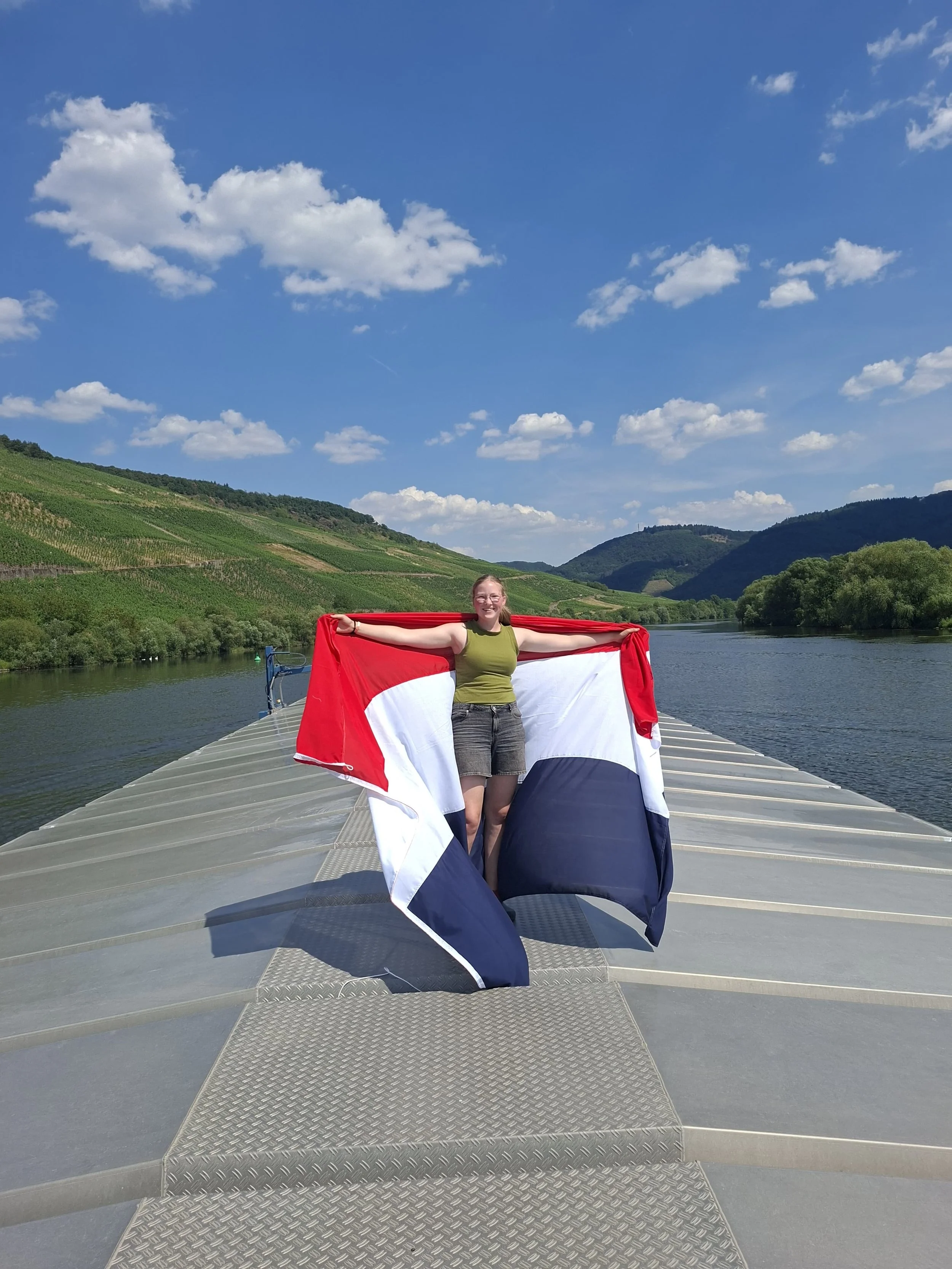 Vrouw staat op een ponton langs een rivier, houdt een Nederlandse vlag uitgestrekt met beide armen, omringd door groene heuvels en een blauwe lucht met wolken.