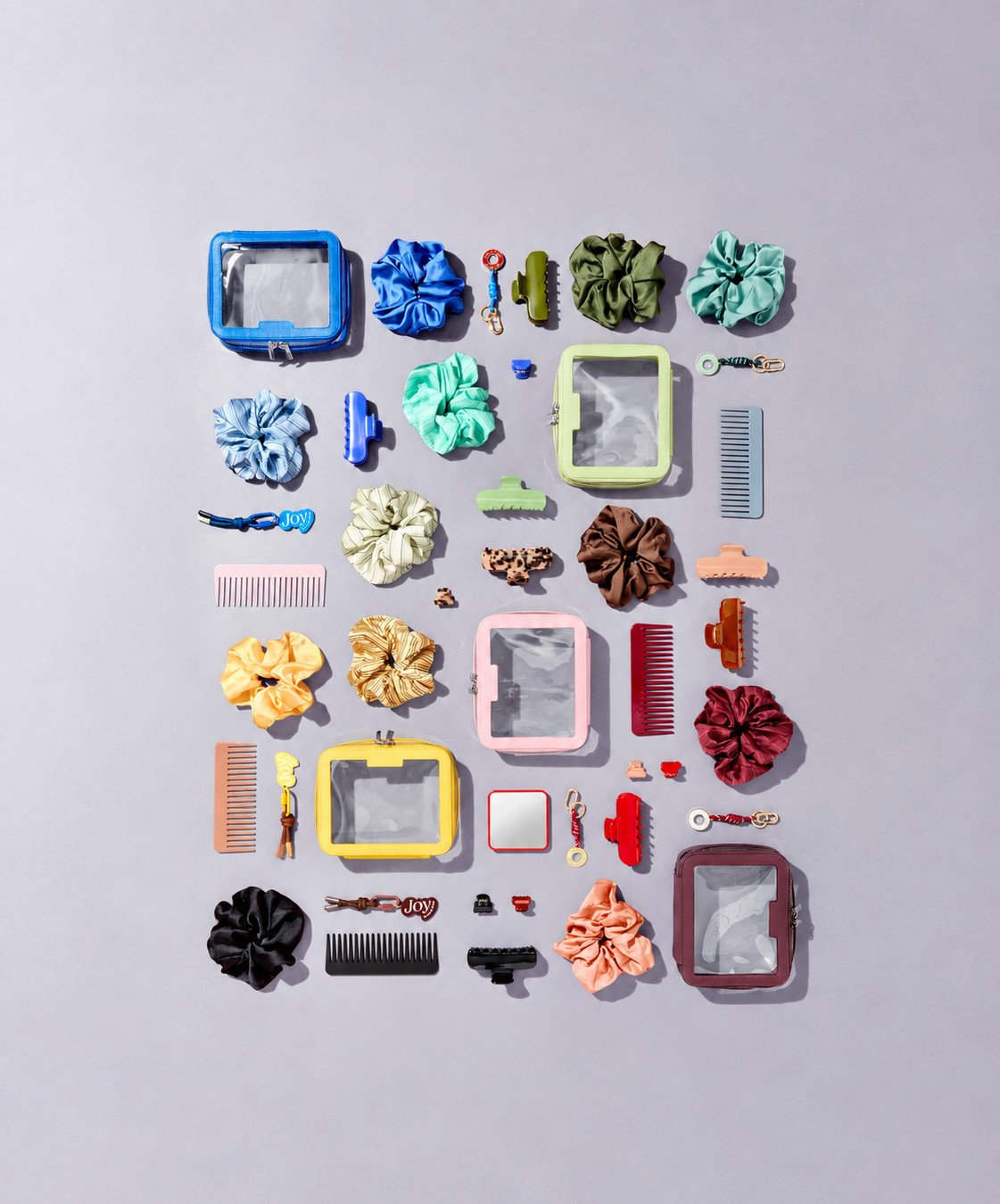 Colourful flatlay of wash bags, scrunchies, hair clips and pocket mirrors in blue, green, yellow, pink, red, brown and black against a purple background