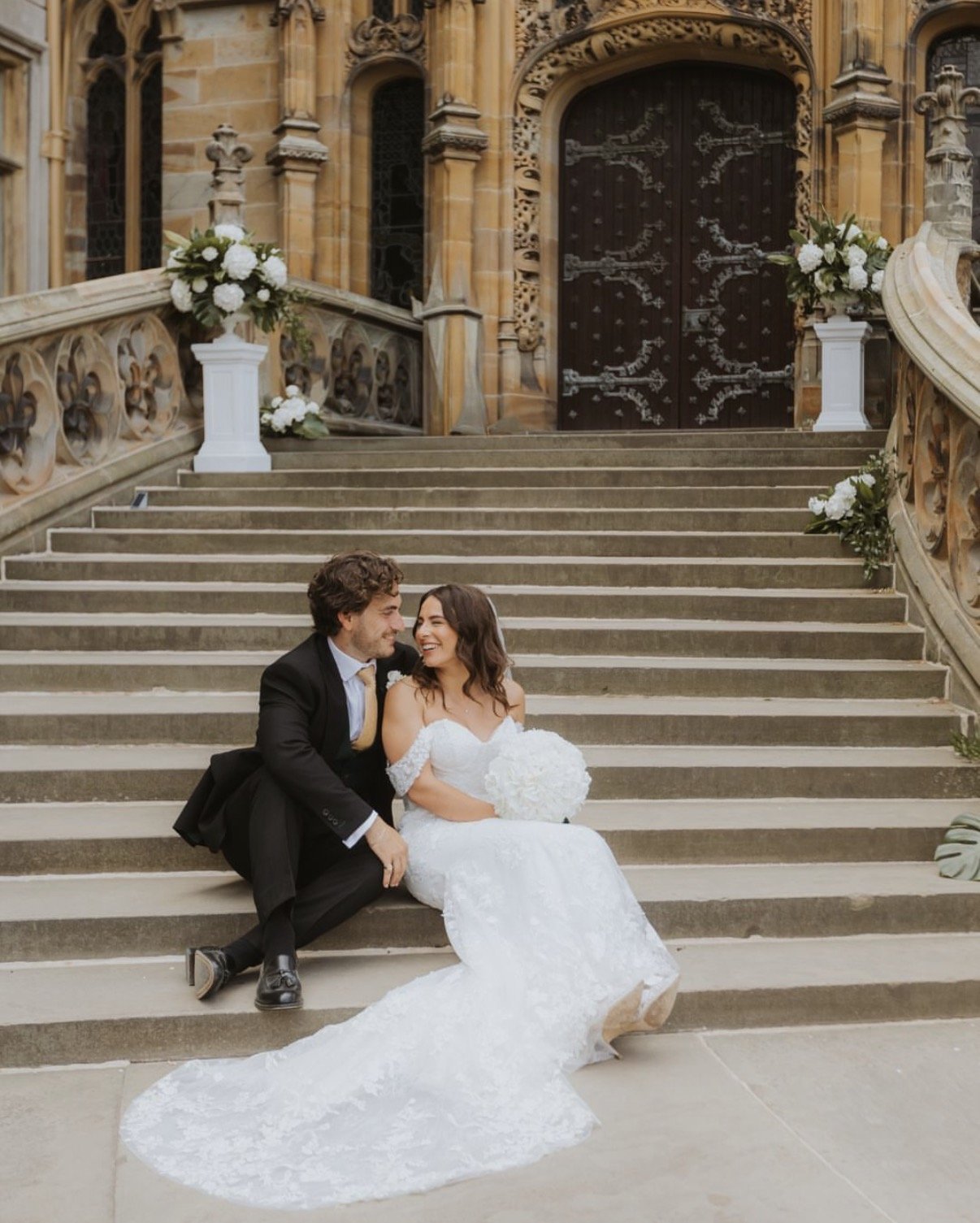 A newlywed couple sitting on the steps outside a historic building with Gothic architecture, dressed in wedding attire, smiling and looking at each other. The bride is holding a bouquet of white flowers, and the scene is decorated with large white fl