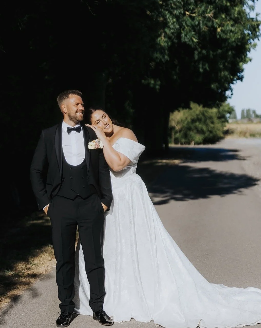 A bride and groom stand together outdoors on a sunny day, smiling and sharing a joyful moment after their wedding. The groom wears a black tuxedo with a bow tie, and the bride wears a white off-the-shoulder wedding gown.