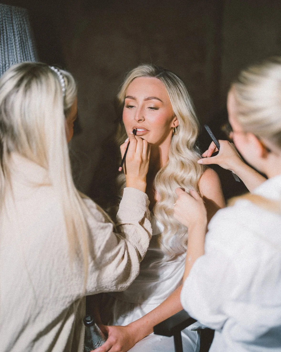 A woman with long blonde hair sitting for a makeup artist to apply lipstick while two makeup artists work on her.
