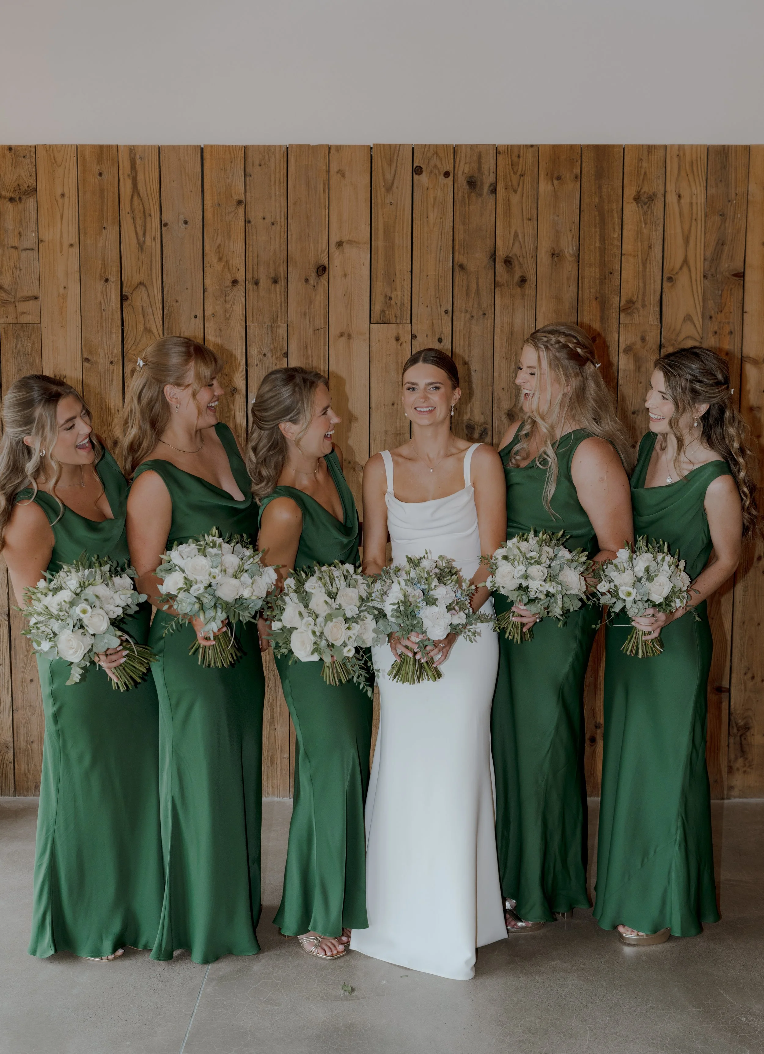 Bride in a white wedding dress standing with six bridesmaids in green dresses, all holding white bouquets, against a wooden wall.
