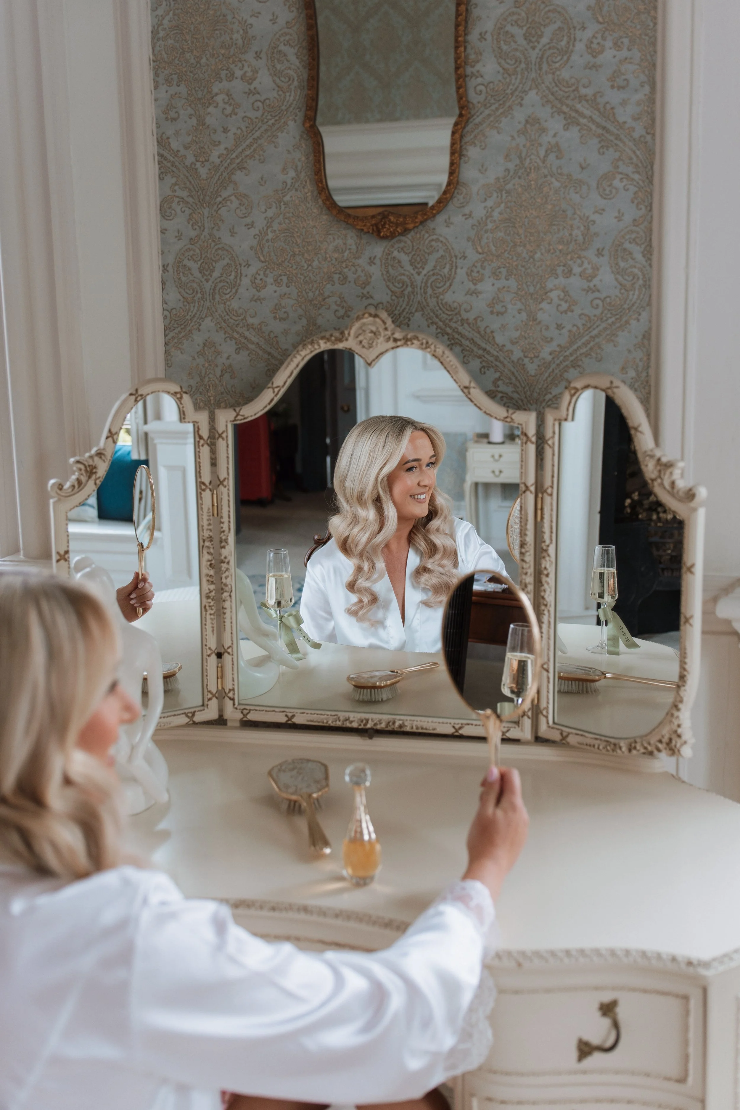 A woman with long blonde hair and a white satin robe is sitting at a vanity, smiling at her reflection in a large ornate mirror. She is holding a small hand mirror and there are several glasses of champagne on the table. The room has elegant decor wi