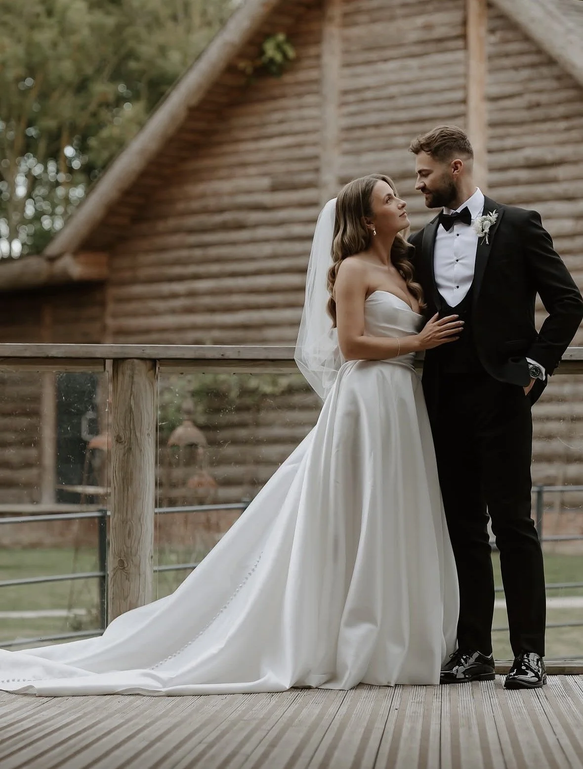 A bride and groom standing on a wooden deck outdoors, with a log cabin in the background. The bride wears a white strapless wedding gown with a long train and veil, and the groom wears a black tuxedo with a bow tie and boutonniere. They look at each 