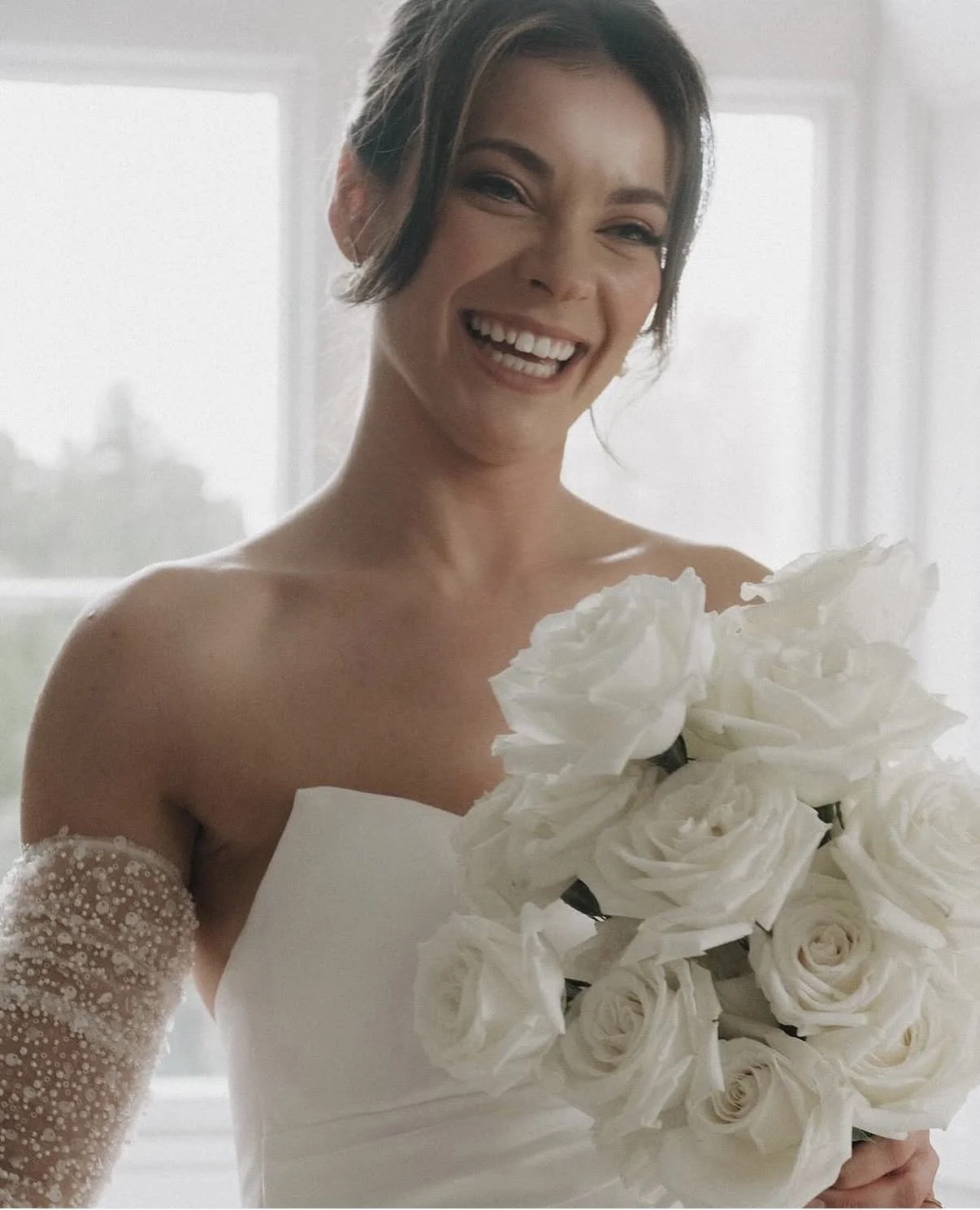 A woman smiling and holding a bouquet of white roses, wearing a strapless wedding dress with a sheer, beaded sleeve on her left arm.