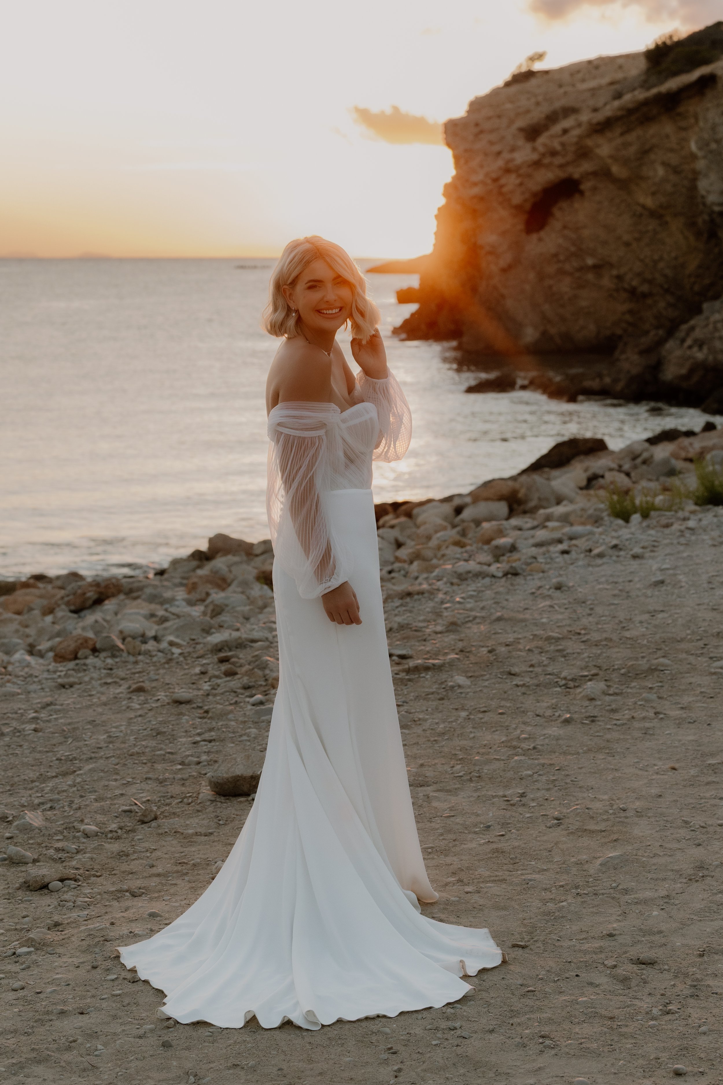 A woman in a white dress standing on a rocky beach at sunset, smiling and looking at the camera with a large rock formation in the background.