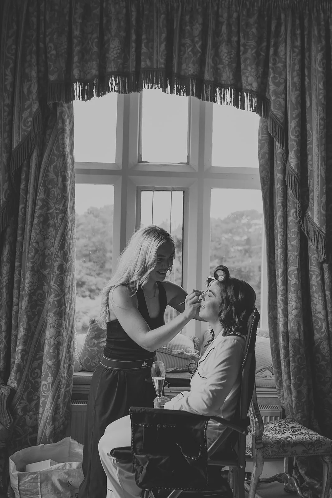 A woman in a white outfit sitting in a chair as another woman applies makeup to her face, holding a makeup brush. They are in a room with a large window and patterned curtains, and the woman receiving makeup has her eyes closed enjoying the process.