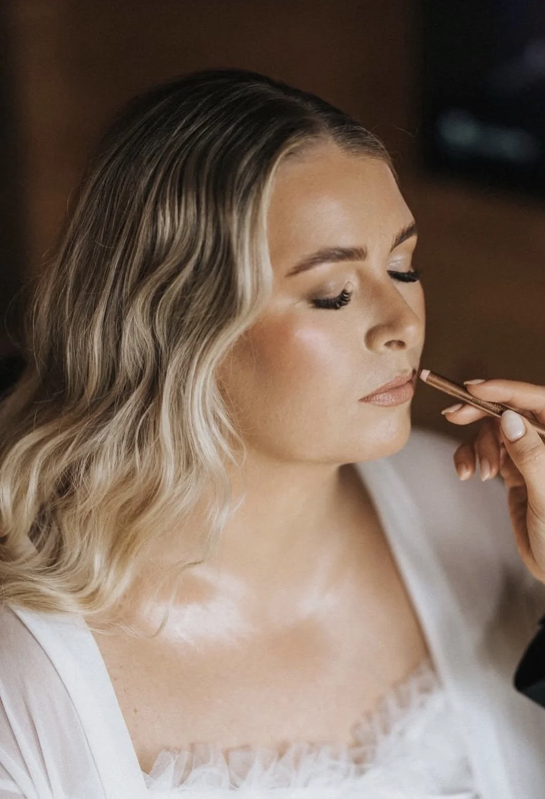 A woman with blonde wavy hair having makeup applied to her lips by a makeup artist.