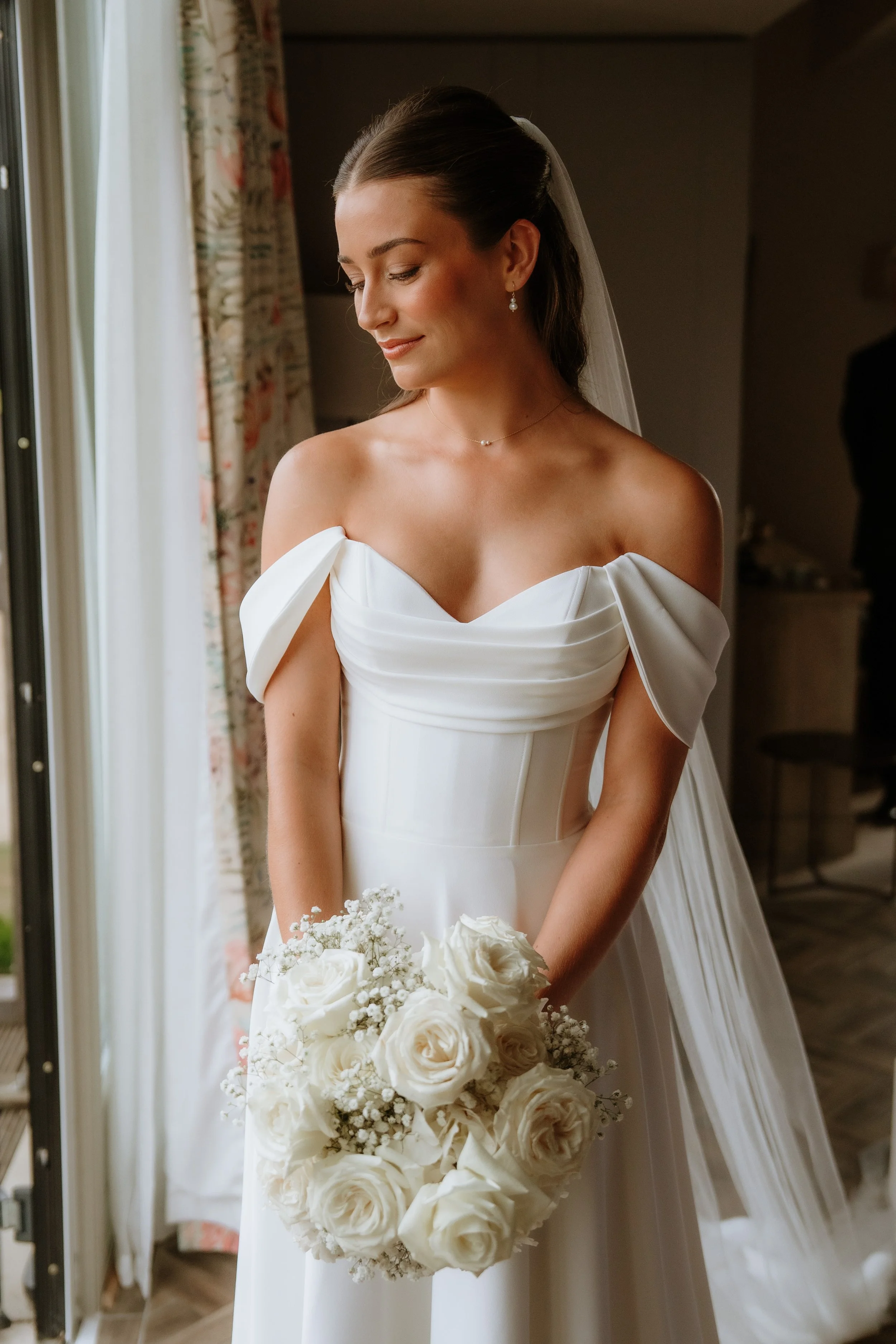 A bride in a white off-the-shoulder wedding gown holding a bouquet of white roses and baby's breath, standing indoors near a window with floral curtains, with a soft smile and looking down.