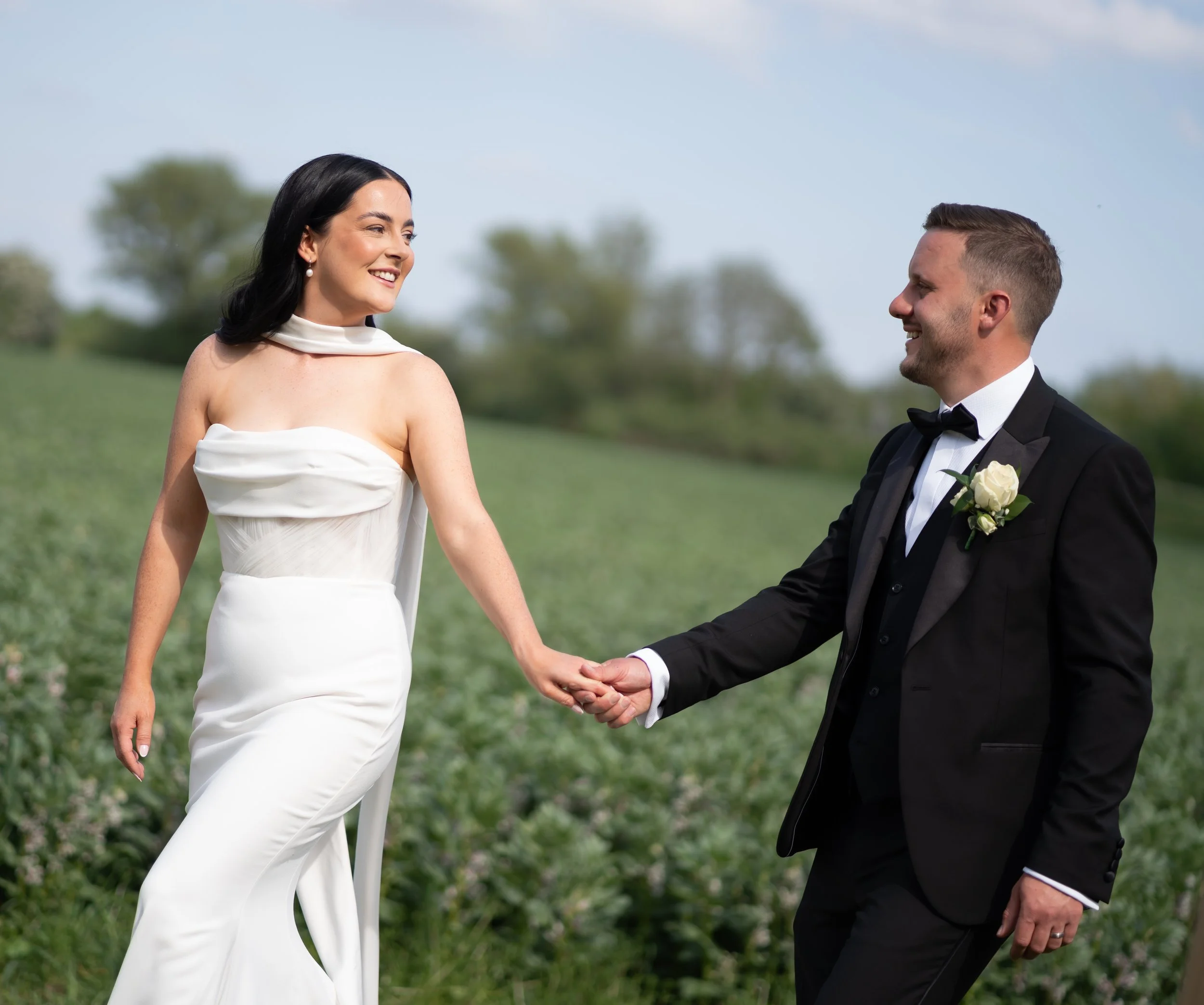 A smiling woman and man holding hands outdoors on a sunny day, with the woman dressed in a white dress and the man in a black tuxedo with a white shirt and black bow tie, in a grassy area with trees in the background.