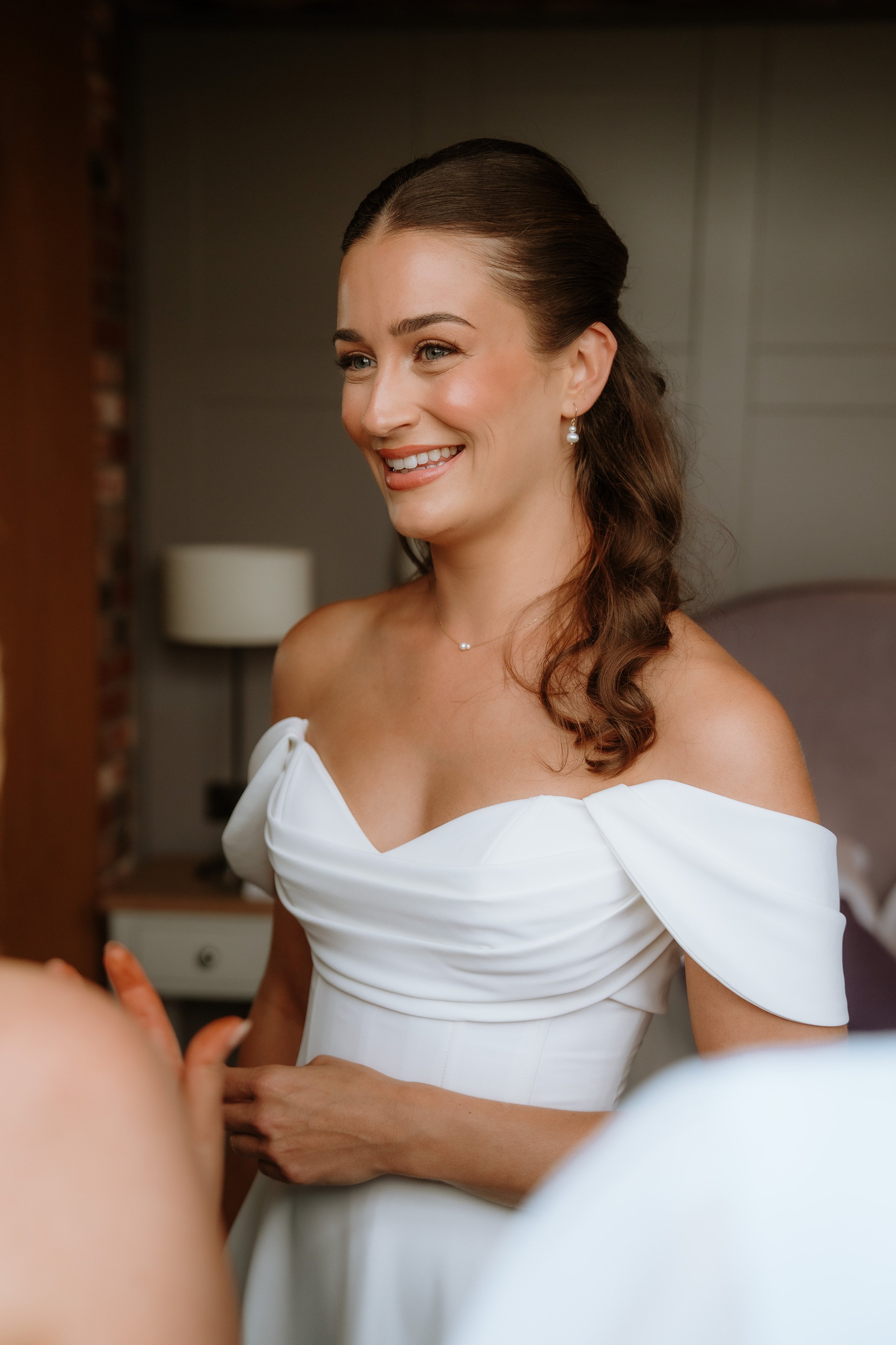 A smiling woman with brown hair styled in a waved half-updo, wearing pearl earrings and a white off-the-shoulder dress, standing indoors.