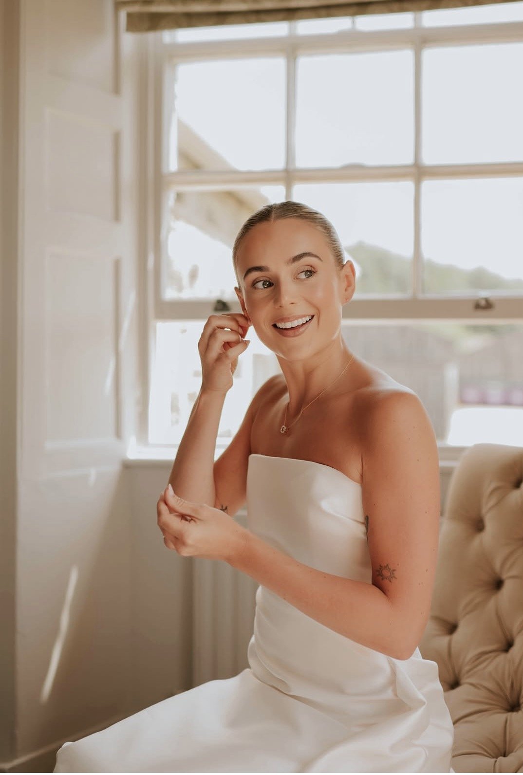 A smiling woman in a white strapless dress sitting on a beige tufted chair, adjusting her earring inside a bright room with large windows.
