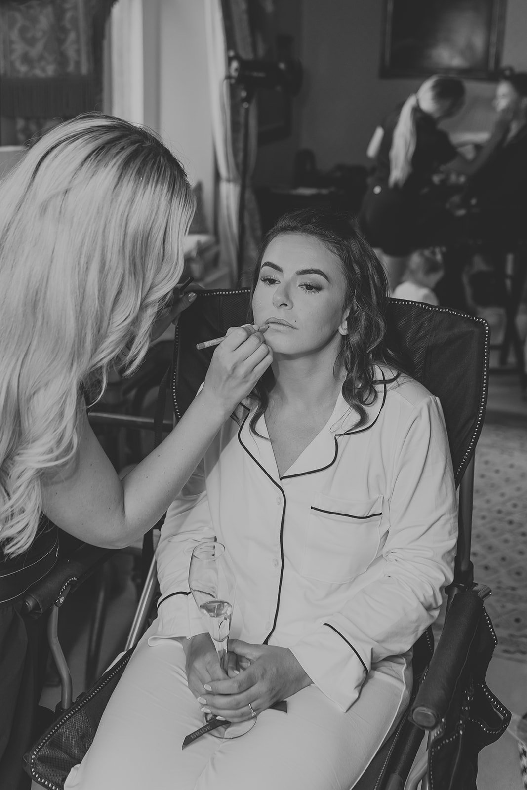 A woman with long dark hair is sitting in a wheelchair wearing pajamas, while another woman with long light hair applies makeup to her face.