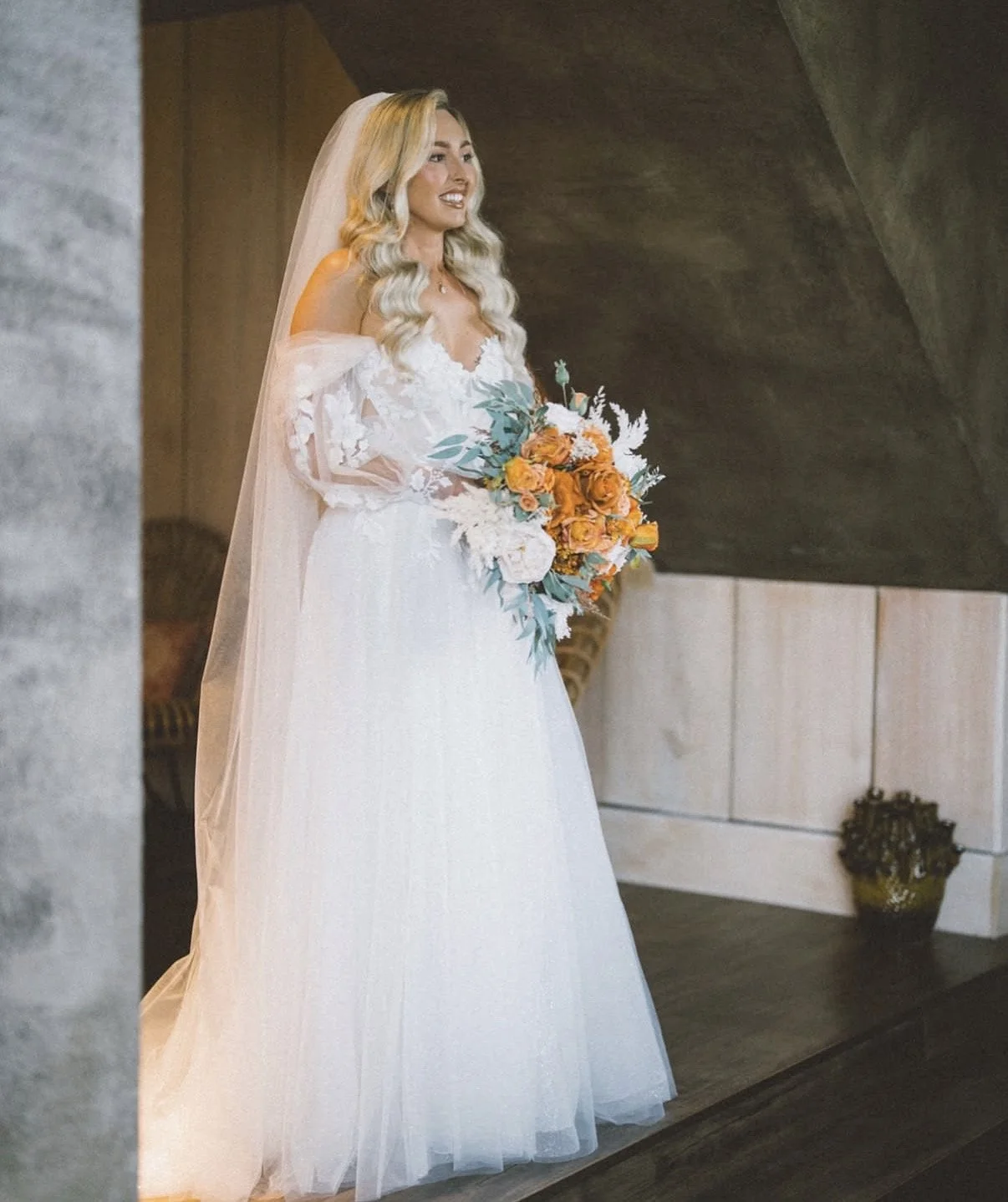 A bride with long wavy blonde hair wearing a white wedding dress and veil, holding a bouquet of orange and white flowers, standing indoors near a charcoal gray wall.
