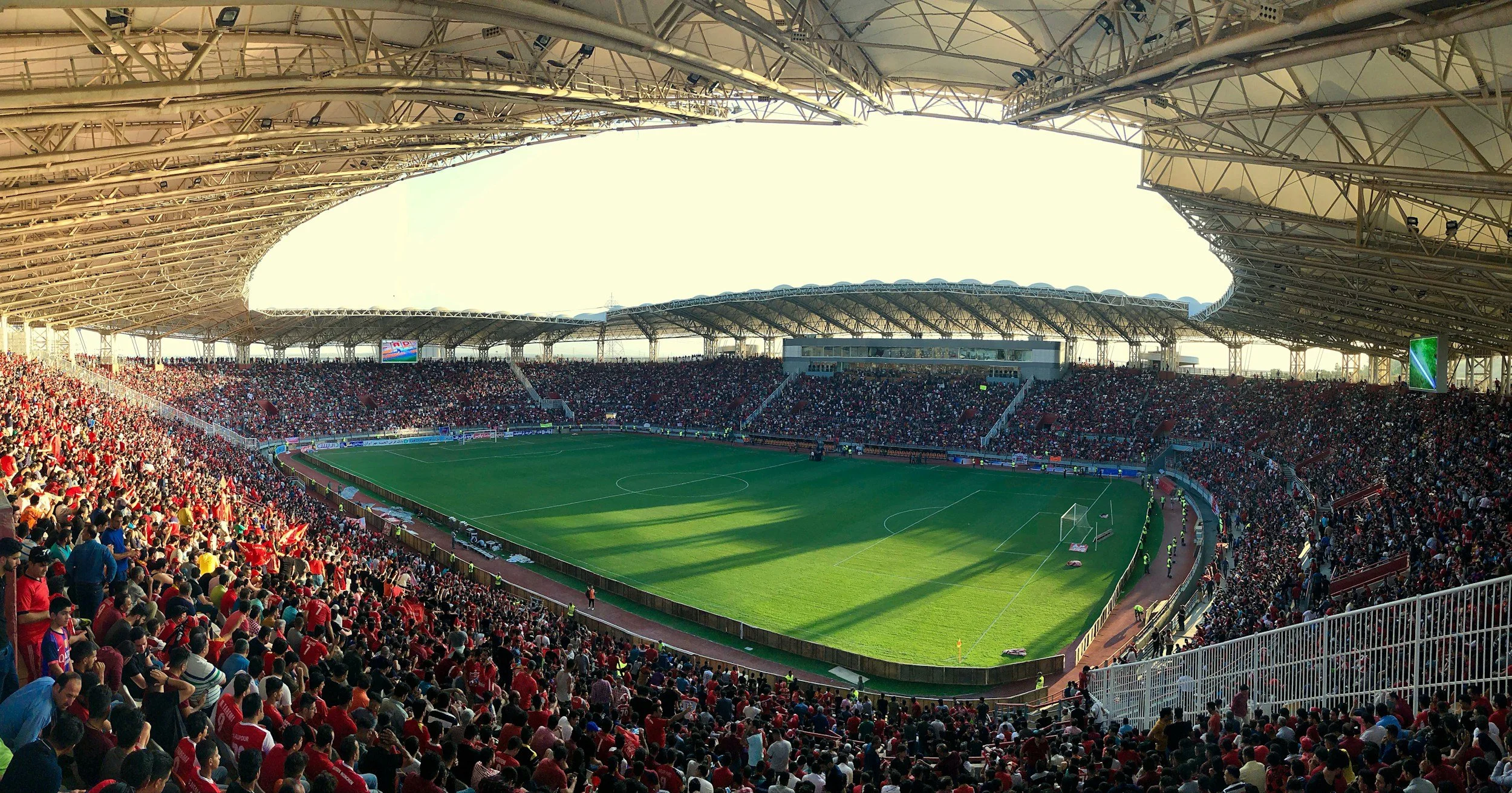 A large crowded football stadium filled with spectators watching a game, with a green field and a roof structure overhead.