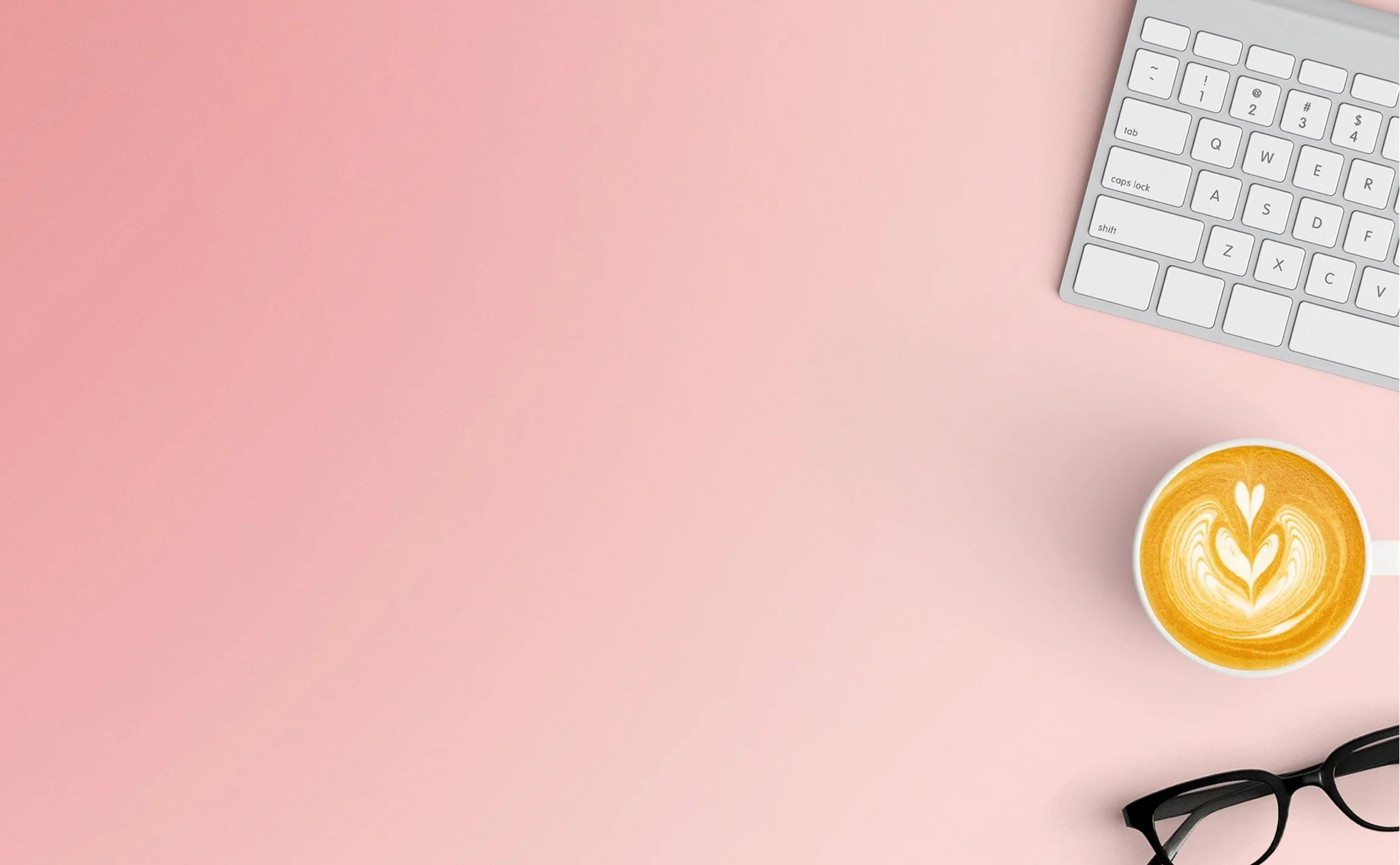 Pink workspace with a white keyboard in the top right corner, black eyeglasses near the bottom right, and a cup of latte with heart-shaped foam art near the bottom right.