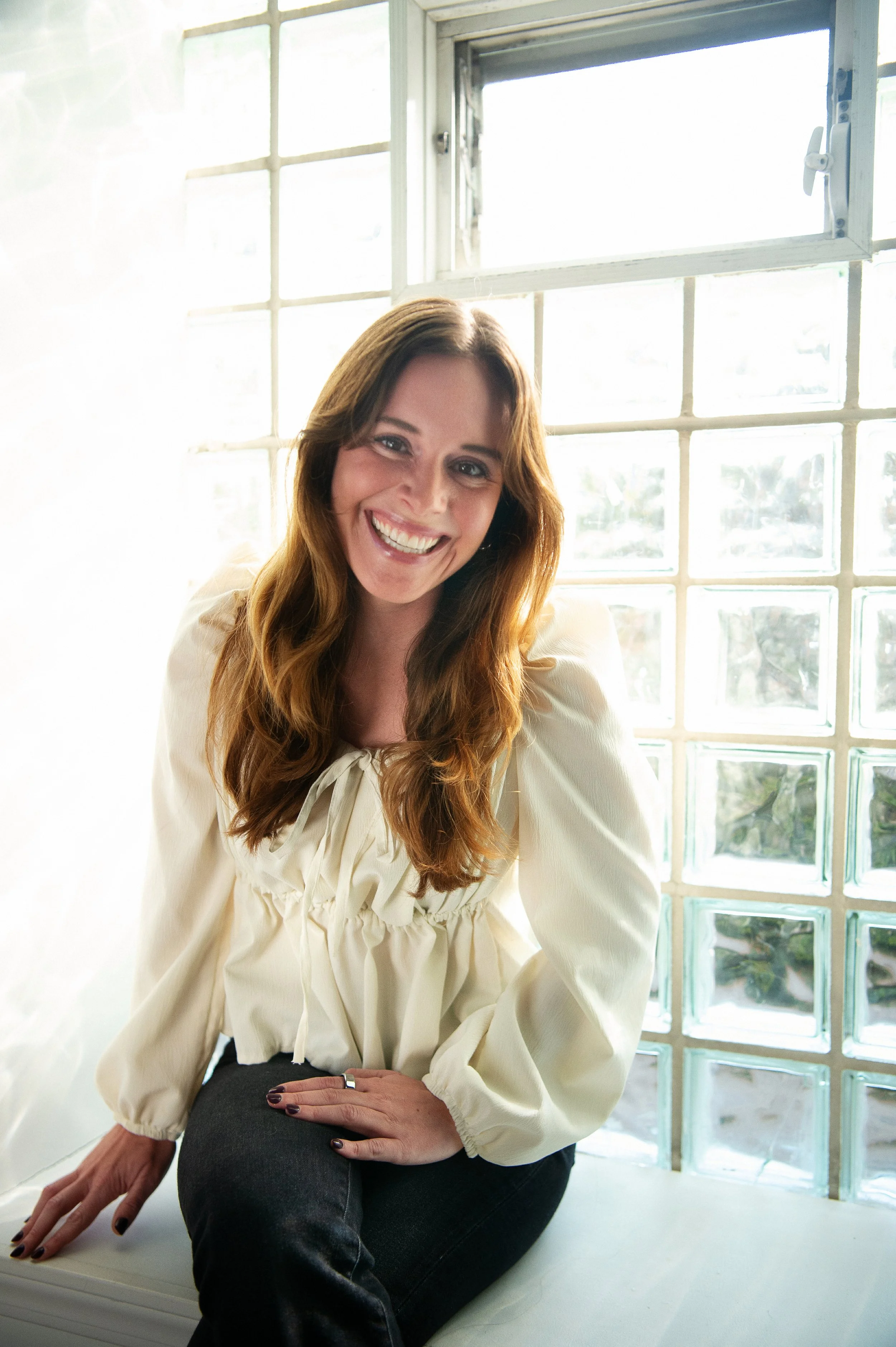 Woman with long hair smiling, seated by a window with glass blocks, wearing a cream-colored blouse and dark jeans.