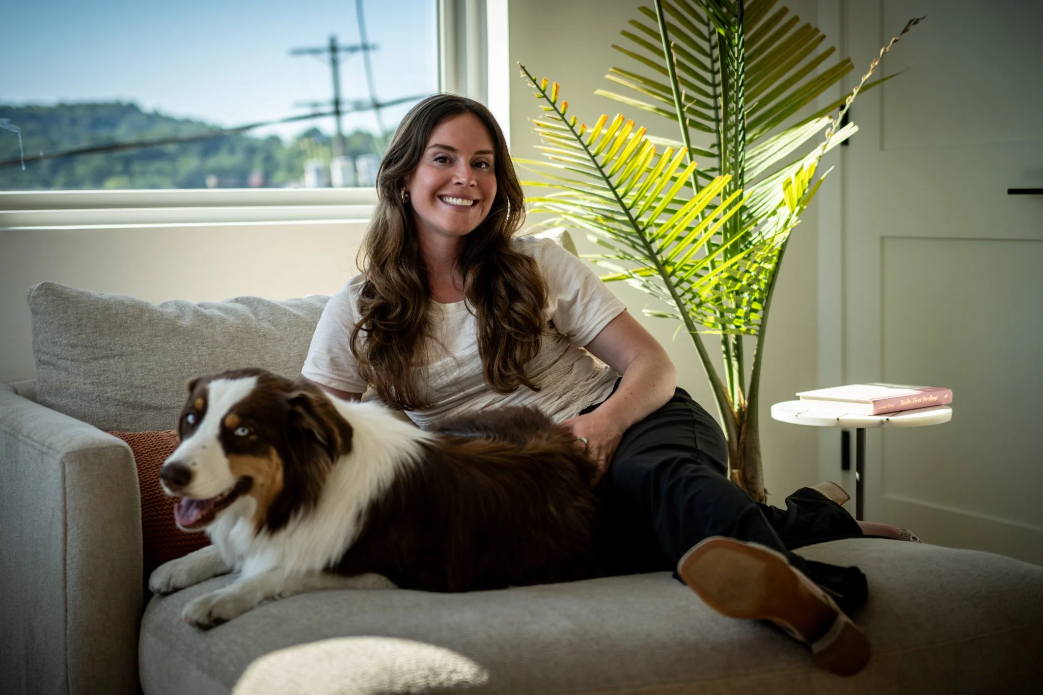 A woman with long brown hair smiling while sitting on a sofa with a dog, in a room with a large window, a green plant, and a small side table with books.