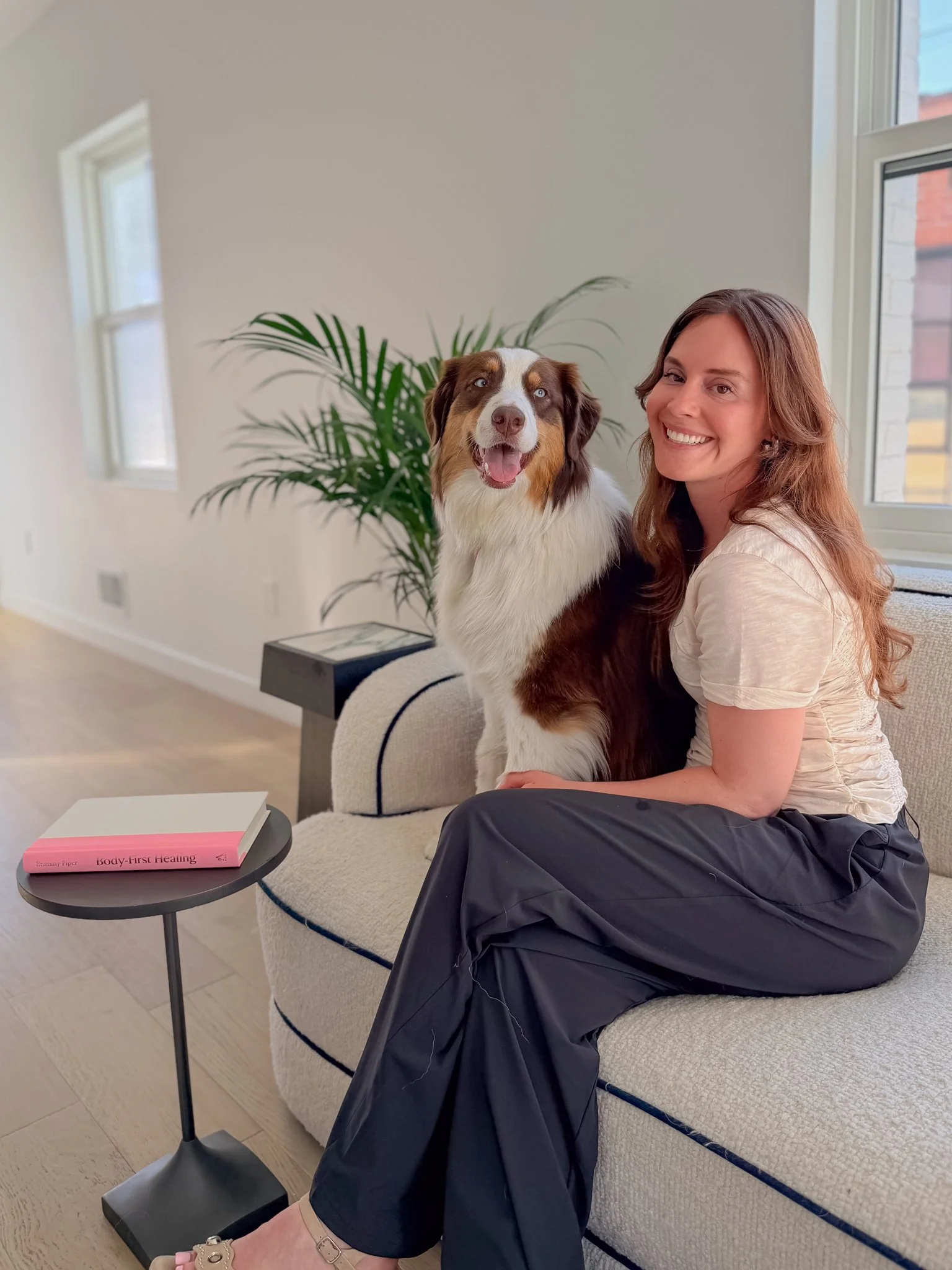 A woman sitting on a beige couch, smiling, with an Australian Shepherd dog, in front of a window with natural light, a green plant, and a pink book titled "Body-First Healing" on a small black round table.