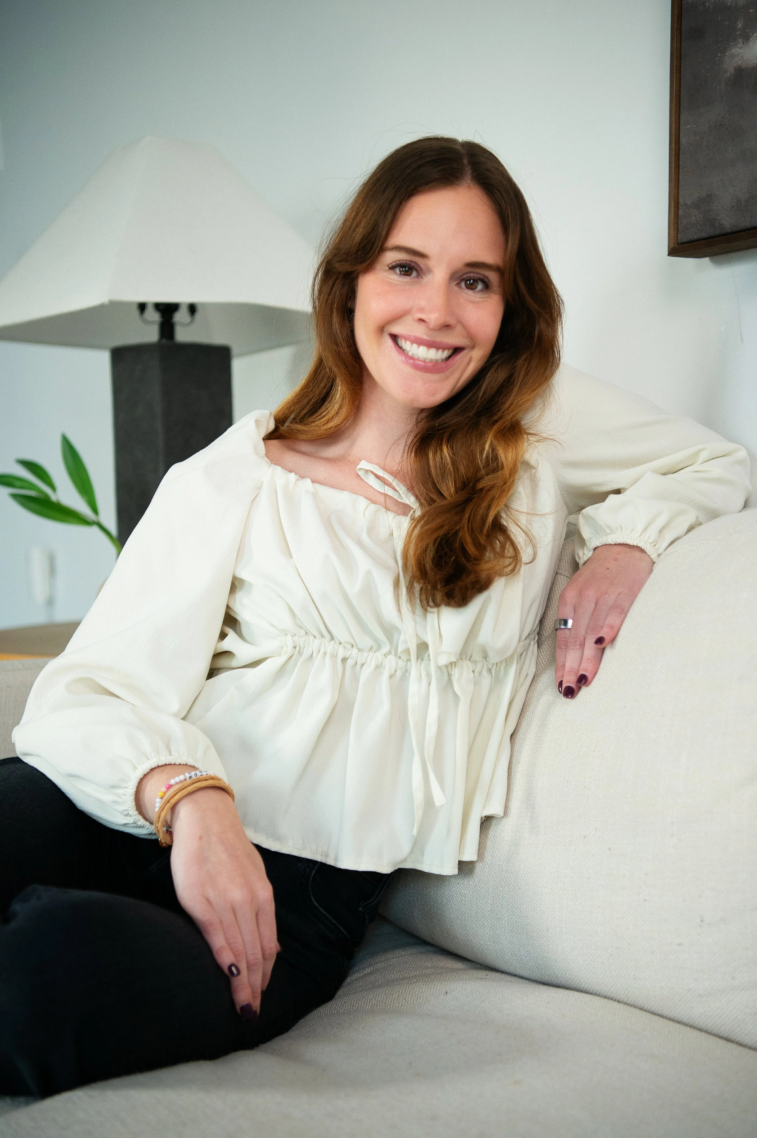 A woman with long wavy reddish-brown hair, smiling, sitting on a beige couch in a cozy living room. She is wearing a white blouse with puffed sleeves, layered bracelets on her wrist, and has dark nail polish. A lamp with a white shade, a small green plant, and a framed artwork are visible in the background.