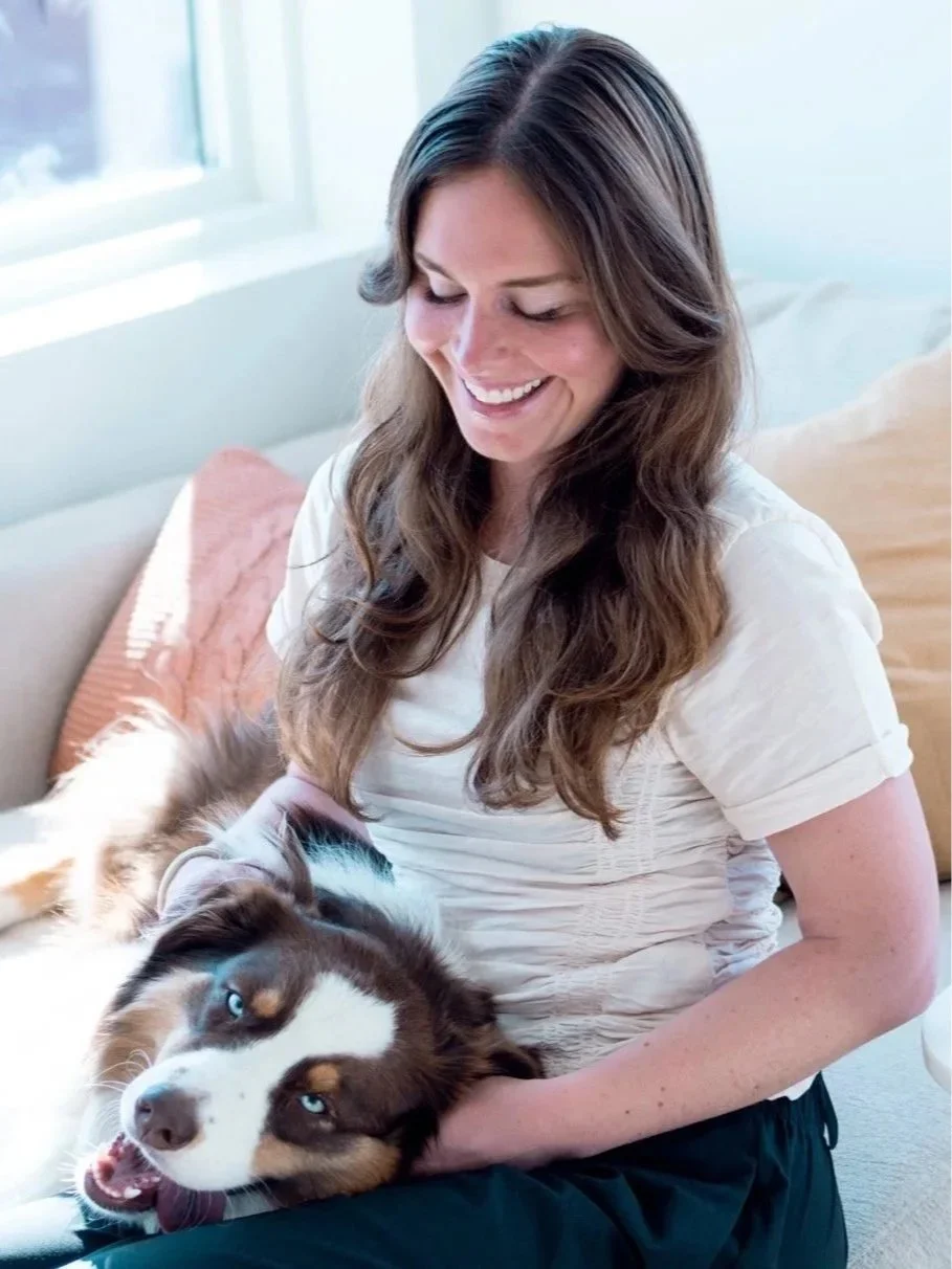 A woman with long brown hair smiling while sitting on a sofa, petting a tricolor Australian Shepherd dog with blue eyes.
