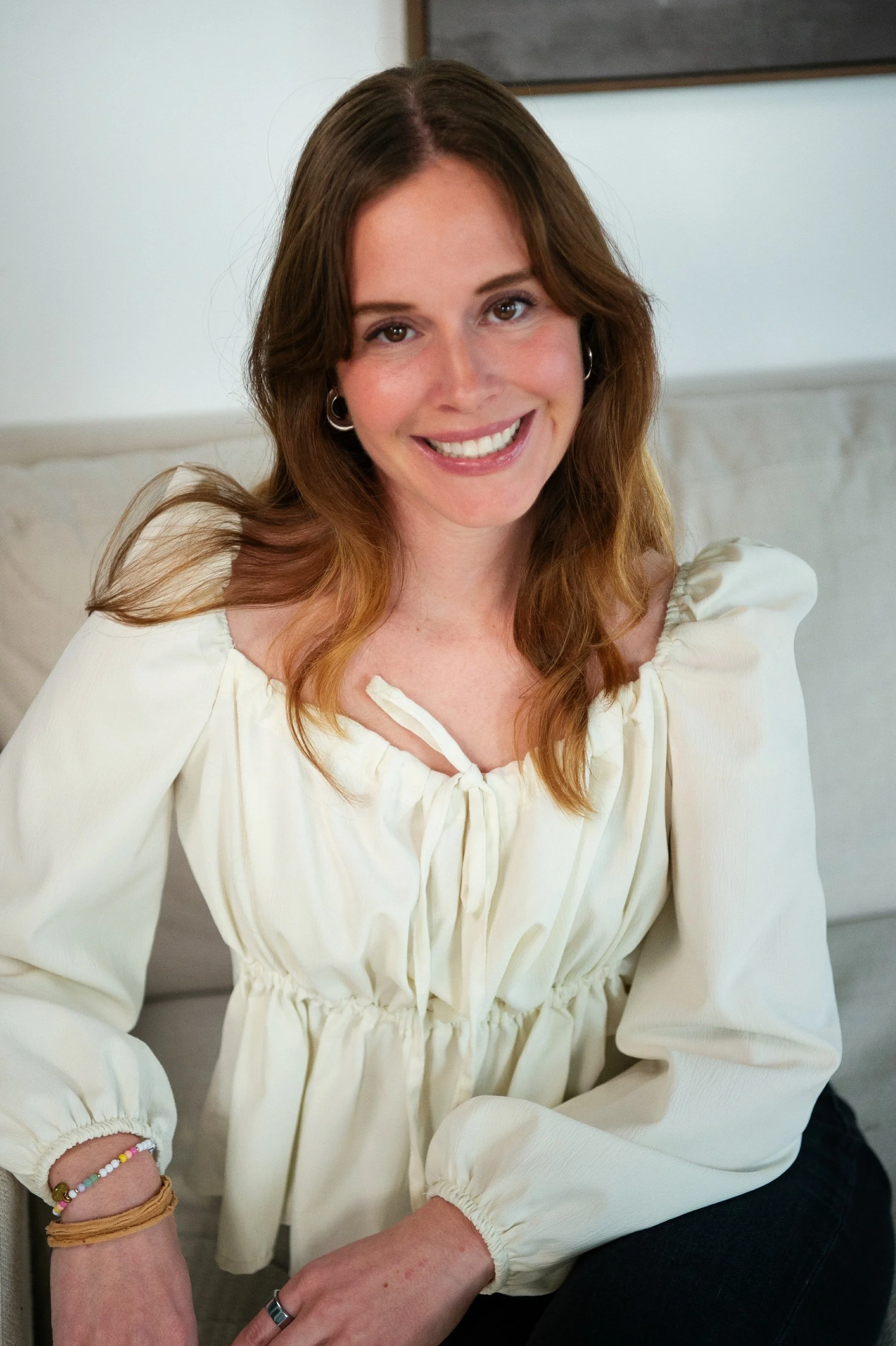 A woman with light brown hair and a bright smile, wearing a cream-colored blouse with puffed long sleeves, sitting indoors in a cozy setting.