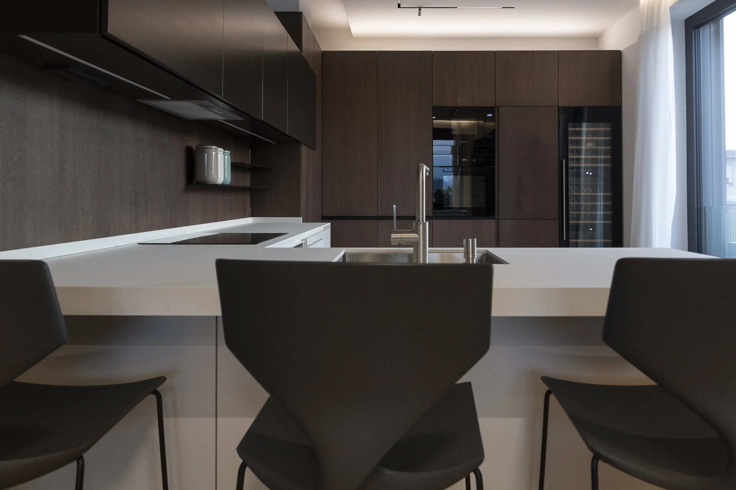 Modern kitchen with dark wooden cabinets, white countertops, a sink with a faucet, and a built-in oven. There are chairs around the countertop, and a large window with sheer white curtains letting in natural light.
