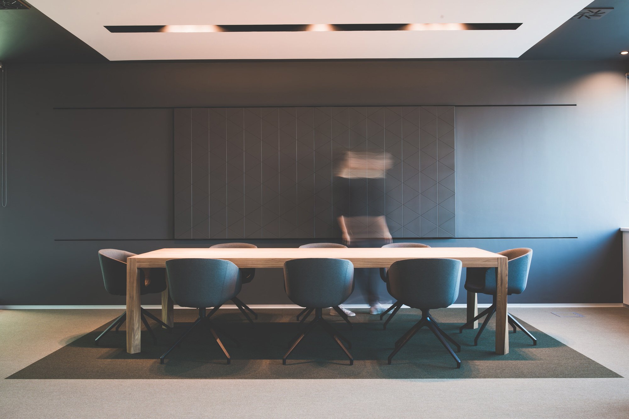 Modern conference room with a long wooden table surrounded by black chairs, a dark grey wall with a geometric patterned panel, and a ceiling with recessed lighting. A blurred reflection of a person can be seen on the wall.