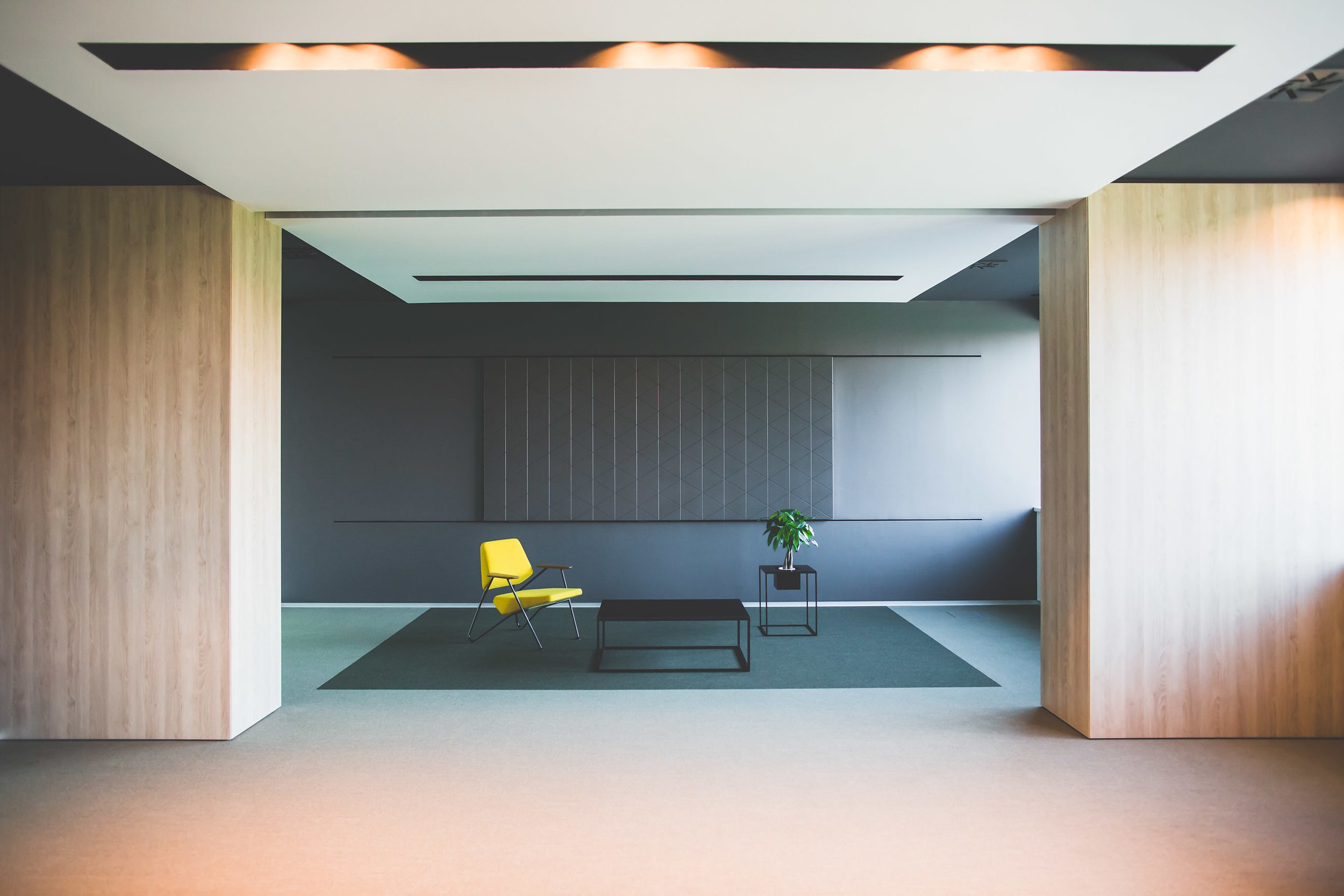Modern interior space with a black chair, a small black side table, a larger black coffee table, and a green plant, framed by wooden columns and a ceiling with recessed lighting.