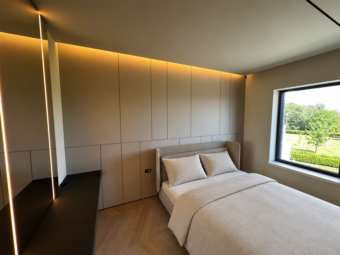 Modern bedroom with beige bed and pillows, large window showing green trees, minimalist wall with warm lighting, black desk-style surface, and wood flooring.