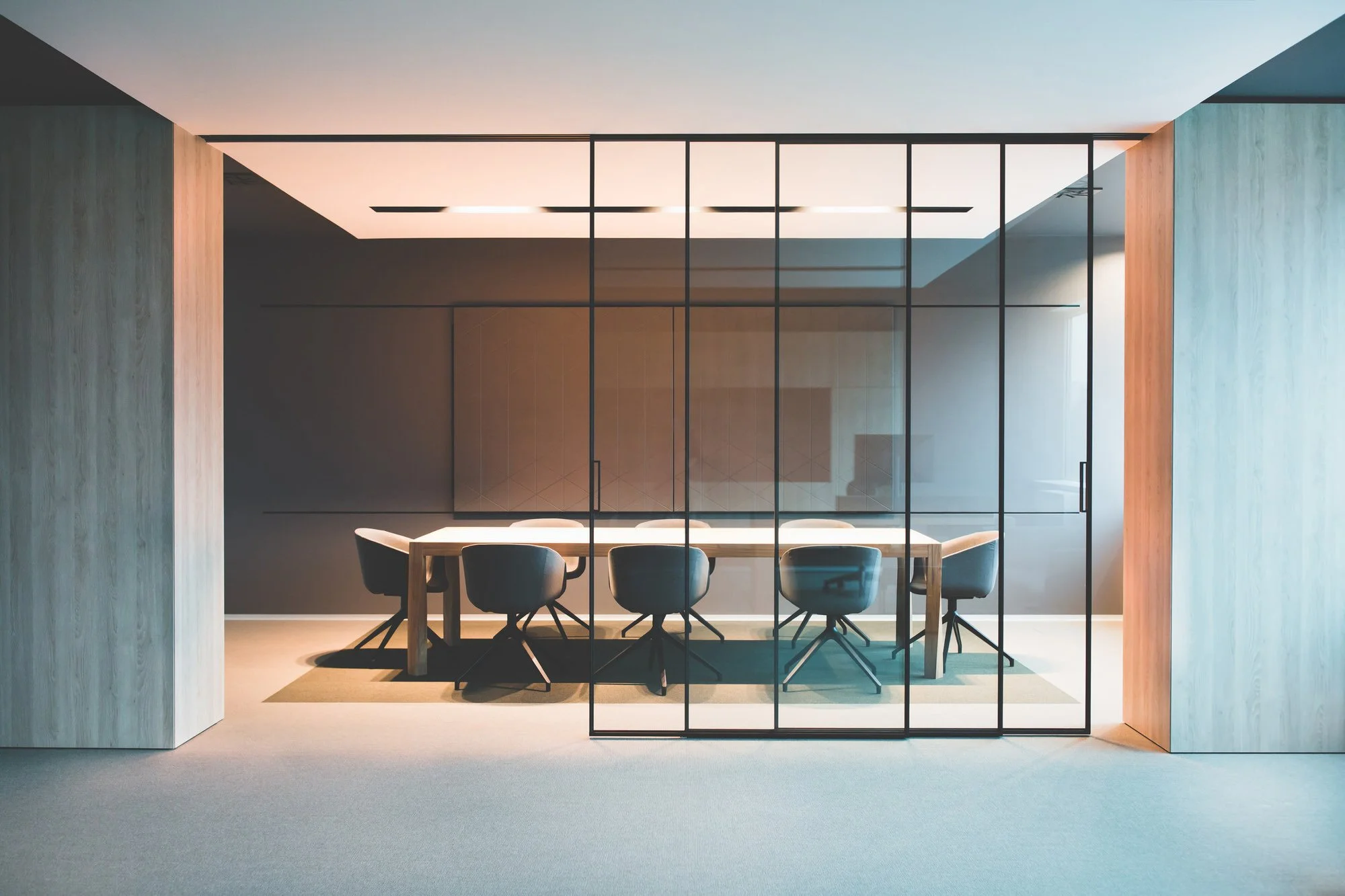 Modern minimalist dining room with a wooden table and six black chairs, separated from the viewer by a black metal framed glass partition, with soft ambient lighting and light-colored walls.
