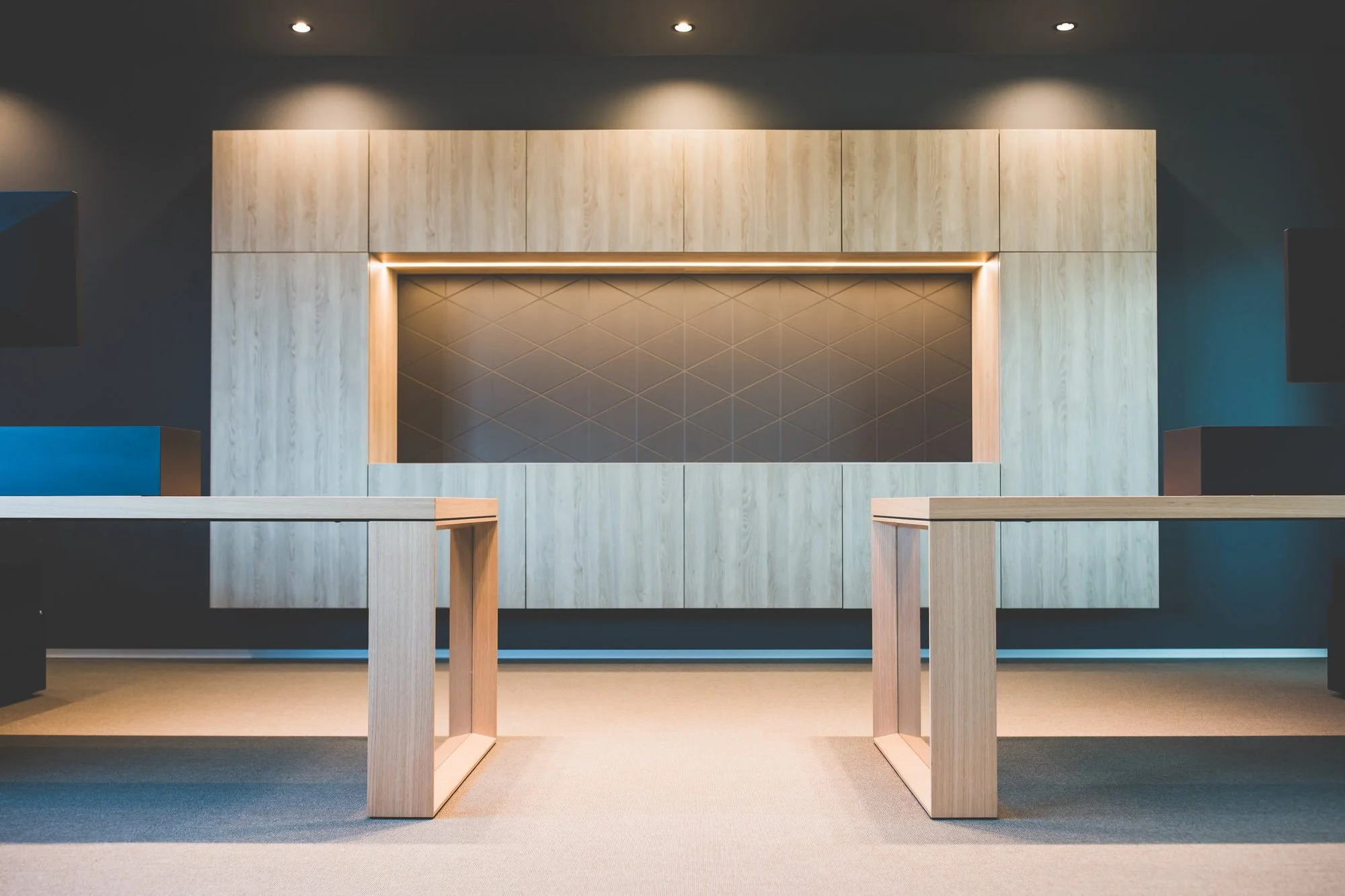 Interior of modern room with a wall featuring a large geometric panel, ceiling with spotlights, and minimalistic wooden tables.