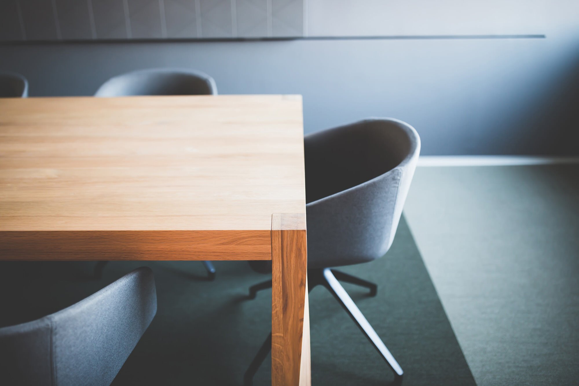 Empty conference room with a wooden table and gray upholstered chairs arranged around it, with a gray wall in the background.