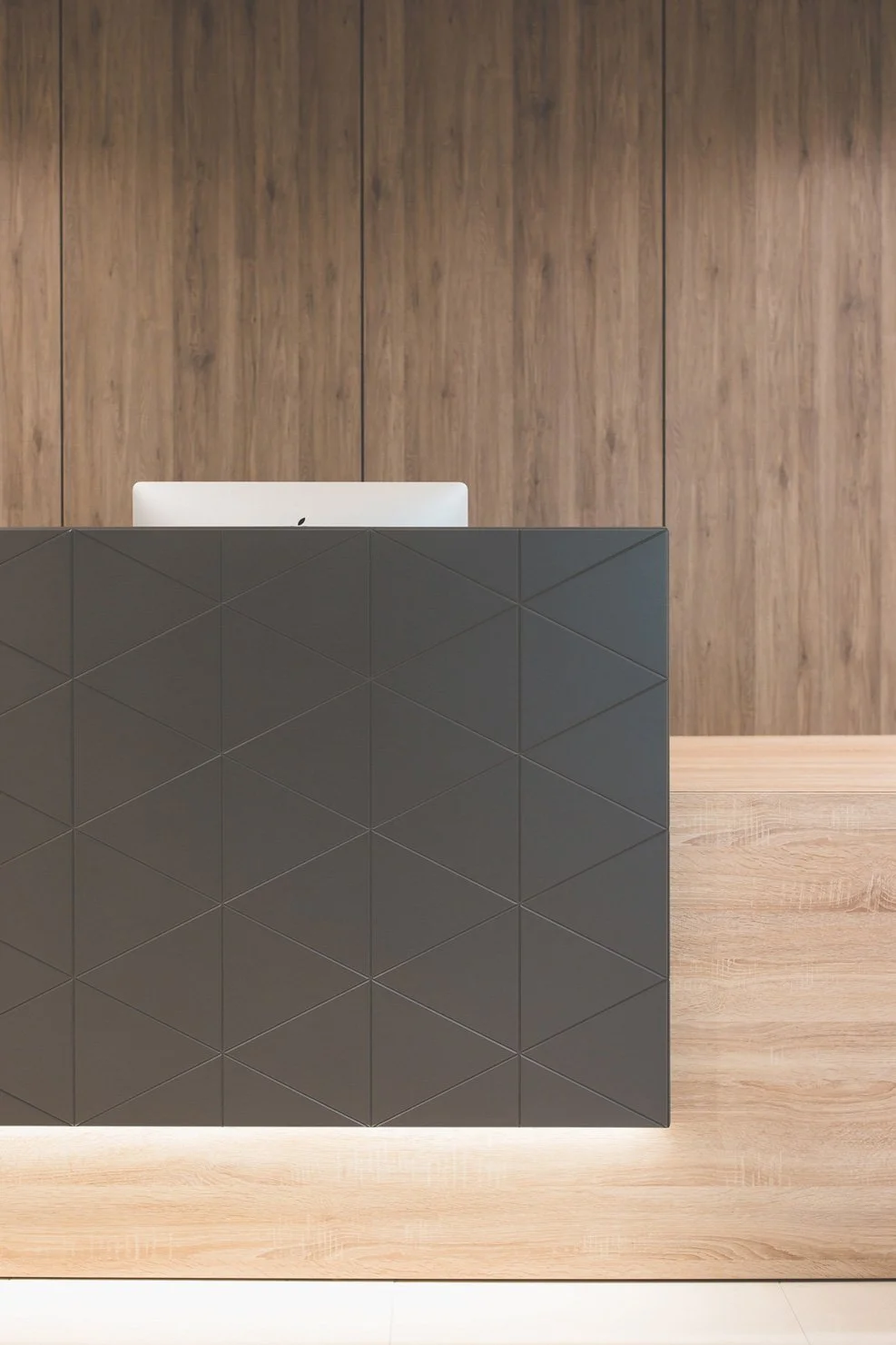 Modern reception desk with laptop, wood-paneled wall background, and light wood flooring.