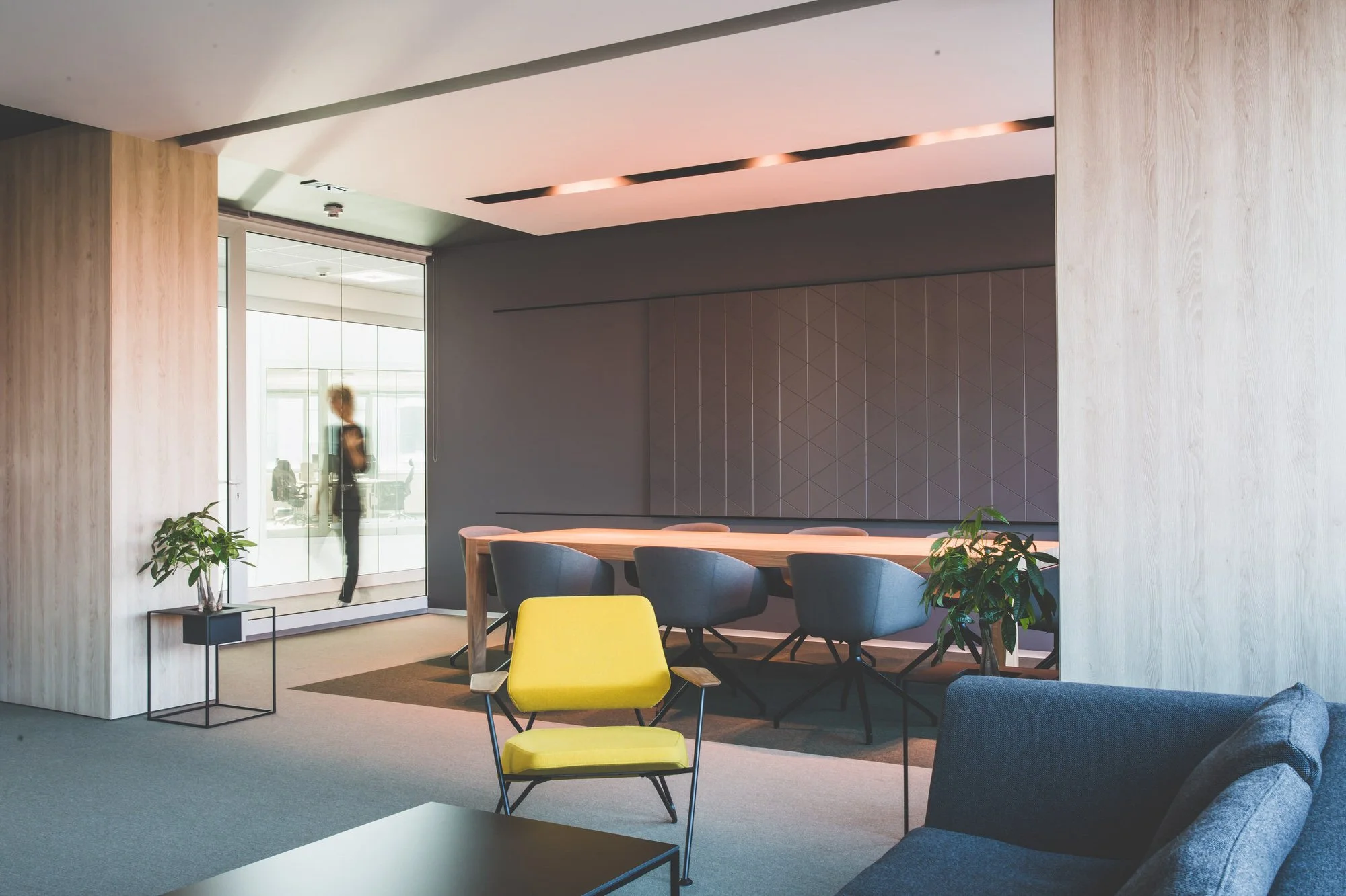 Modern office lounge area with a yellow chair, a gray sofa, a wooden table, and potted plants, with a person visible through glass doors in the background.