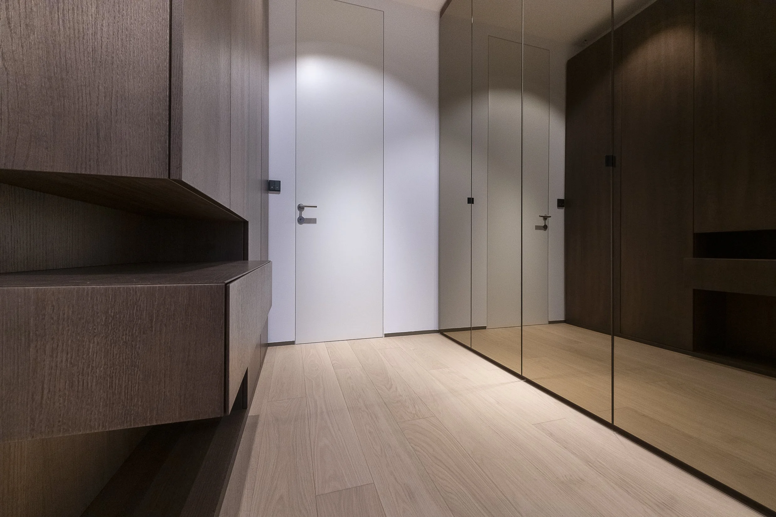 A modern interior hallway with light-colored wood flooring, dark wood and white cabinets along the walls, and a large mirror on the right wall reflecting part of the corridor. There are black door handles and a black light switch on the wall near the white door.