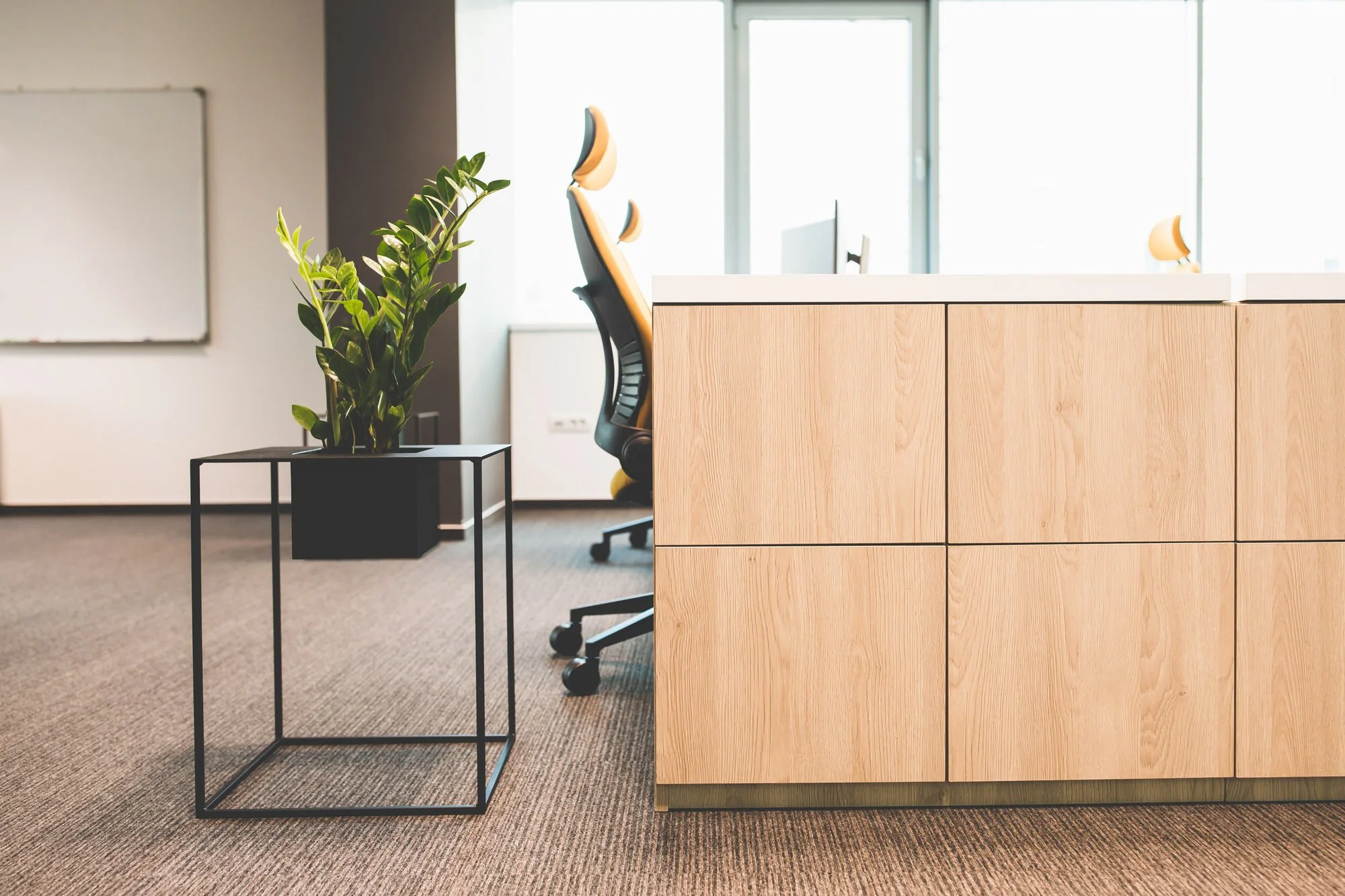 Modern office space with a wooden cabinet, a black metal side table with a green potted plant, an ergonomic office chair, large windows in the background, and a whiteboard on the wall.