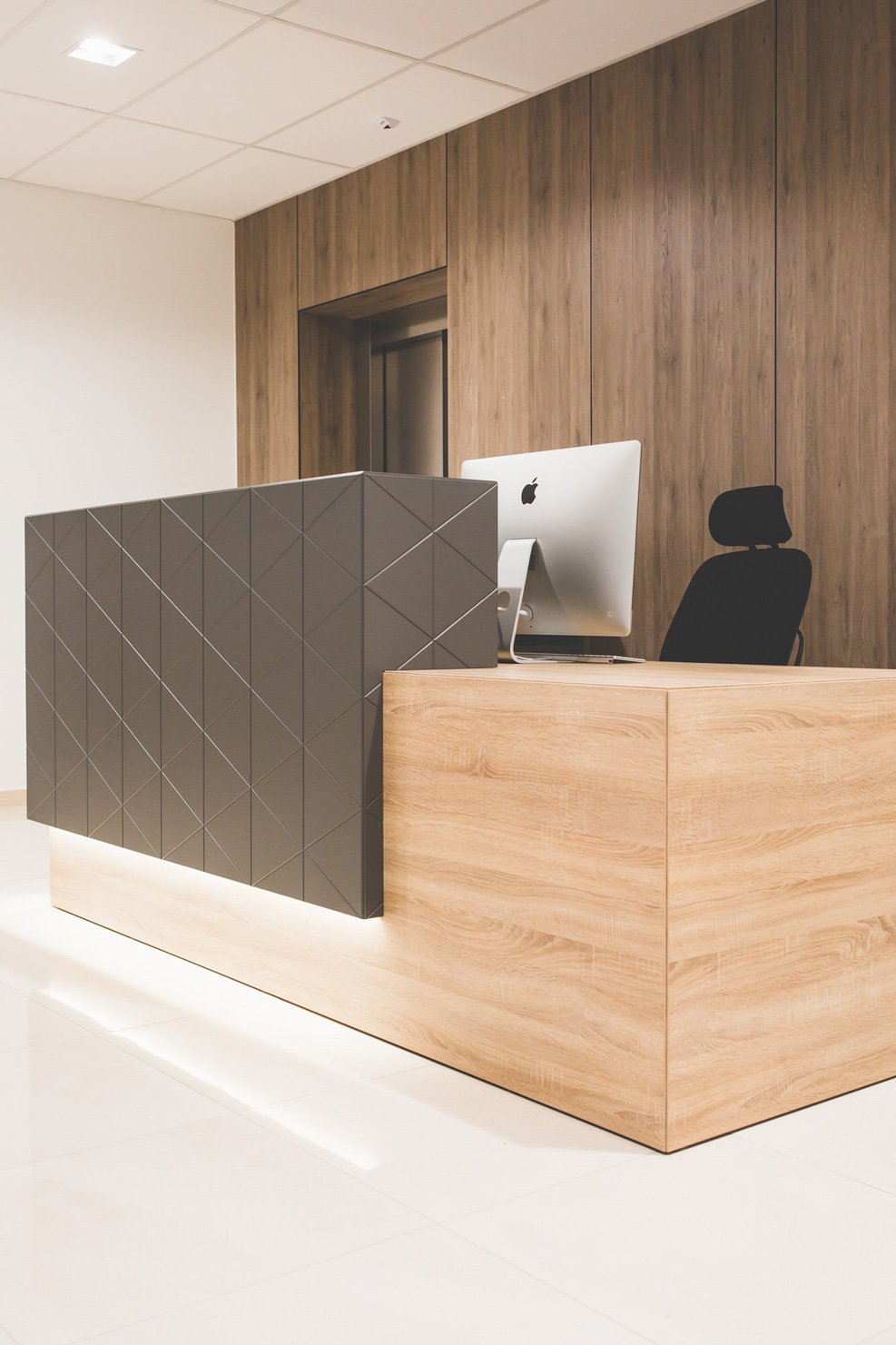 Modern office reception desk with a black geometric front panel, light wood counter, and an iMac computer behind it. Black office chair and wooden panel wall in background.