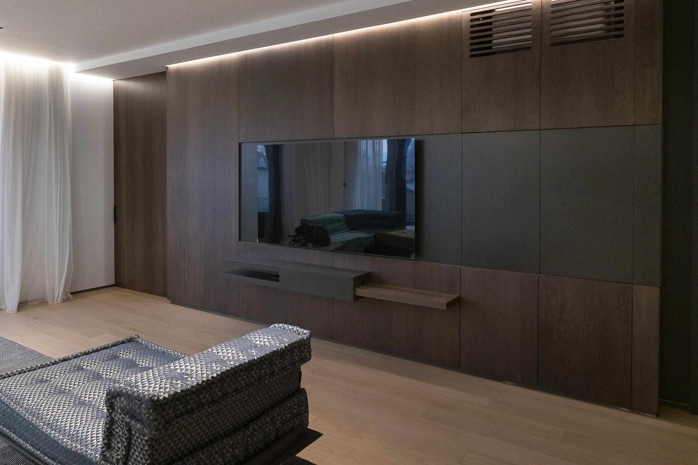 Modern living room with a large flat-screen TV mounted on a wooden panel wall, a navy blue patterned ottoman in the foreground, and beige curtains on the left window.