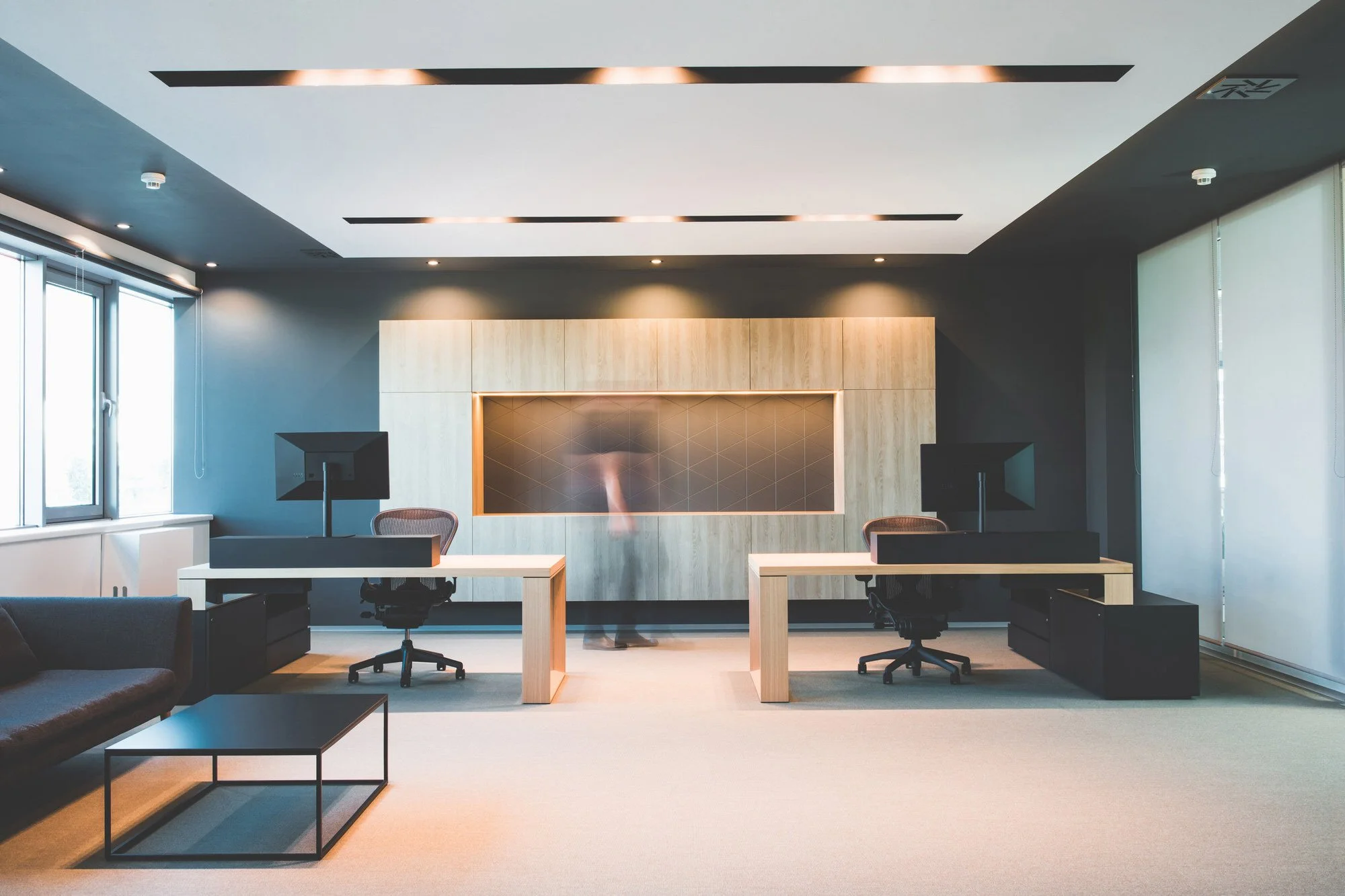 Modern office space with two desks, two office chairs, two computer monitors, a sitting area with a sofa and coffee table, large windows with blinds, and a wall-mounted television with light wood paneling. The ceiling has recessed lighting and air ve