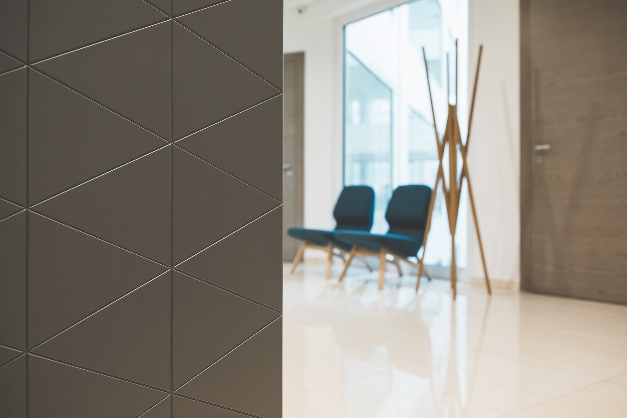 Close-up of a wall with gray tiles arranged in a diagonal pattern, indoors with a large window and two chairs in the background.