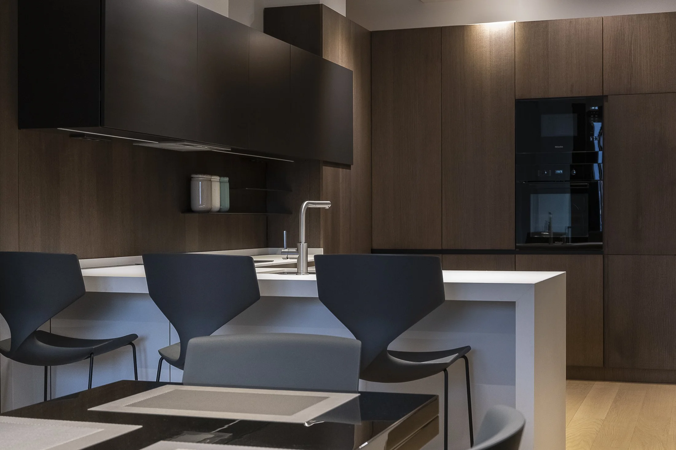 Modern kitchen with dark brown wooden cabinets, white countertop, and black chairs around a white dining table. The kitchen features built-in black appliances and minimal decor, including two jars on a shelf.