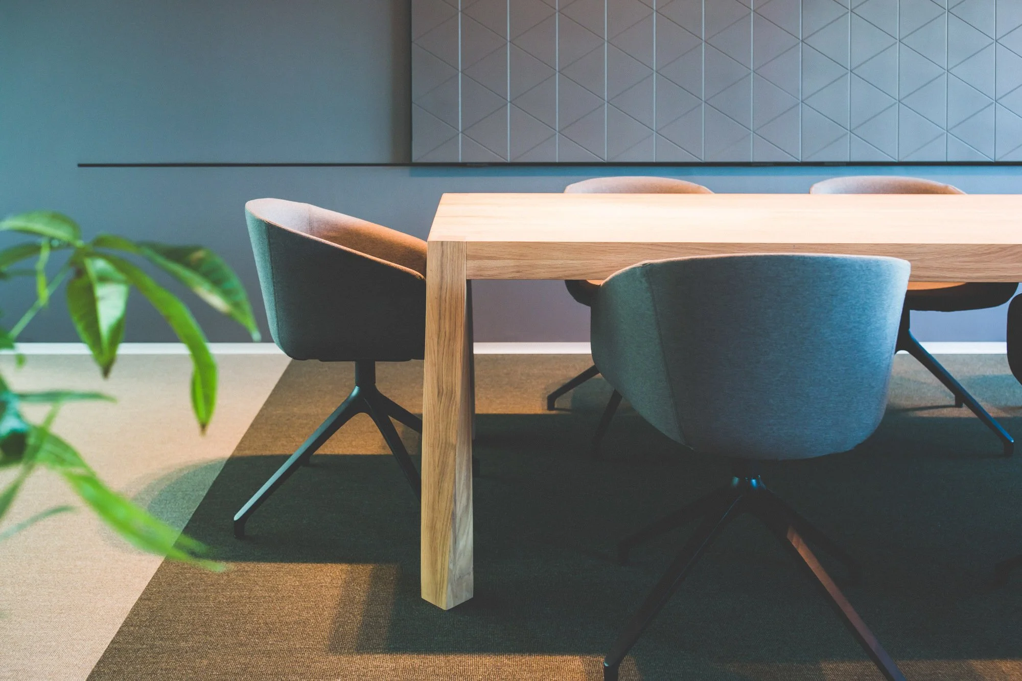 Modern conference room with a wooden table and four upholstered chairs, a plant in the foreground, and a geometric wall design in the background.