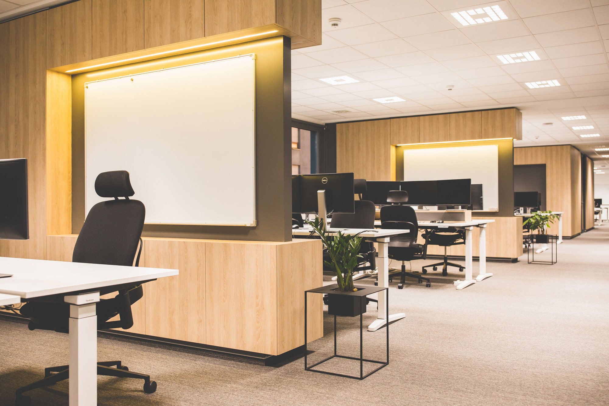 Open office space with work stations, computer monitors, chairs, and plants, featuring wood paneled walls and whiteboards.