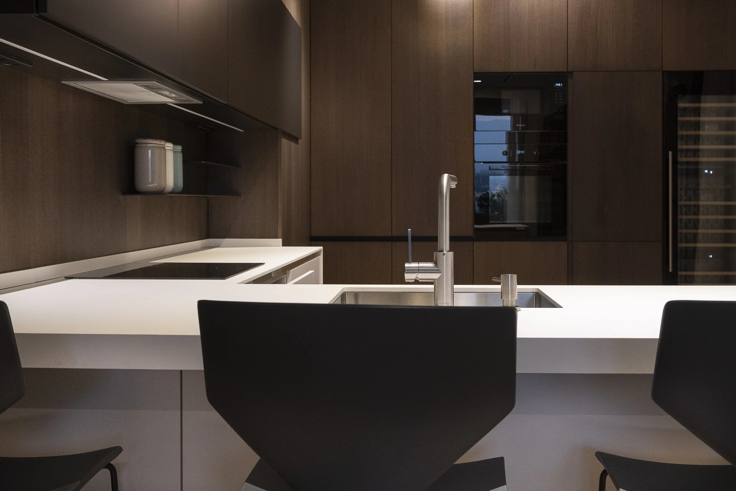 Modern kitchen with white countertop, dark brown cabinets, and black chairs. Features a stainless steel faucet and a built-in oven. A wine fridge is on the right, and a window shows the evening sky.