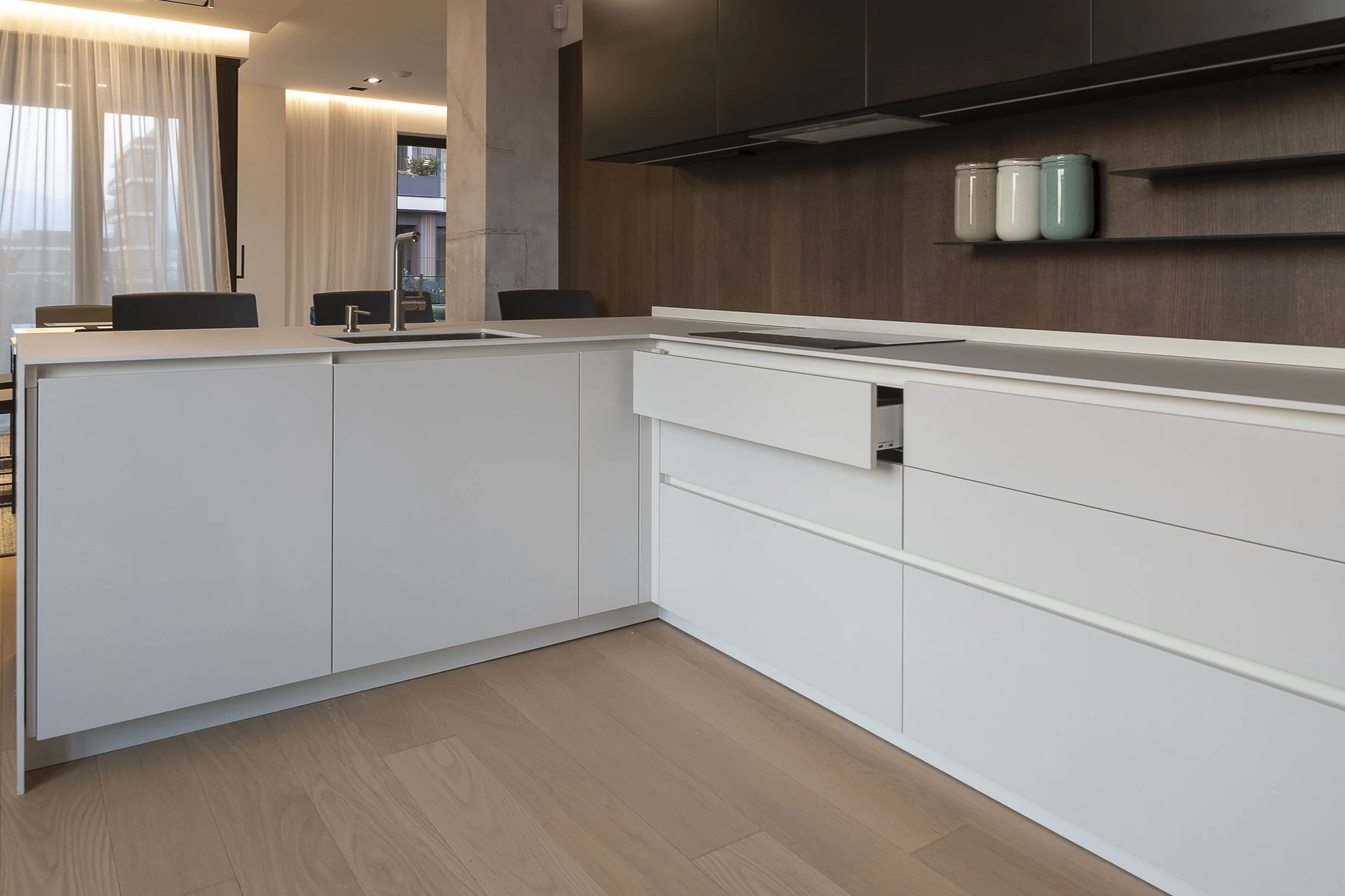 Modern kitchen with white cabinetry, a built-in oven, a stovetop, and a dark wood wall with decorative jars on open shelves. There's a dining area with black chairs and large windows with white curtains letting in natural light.