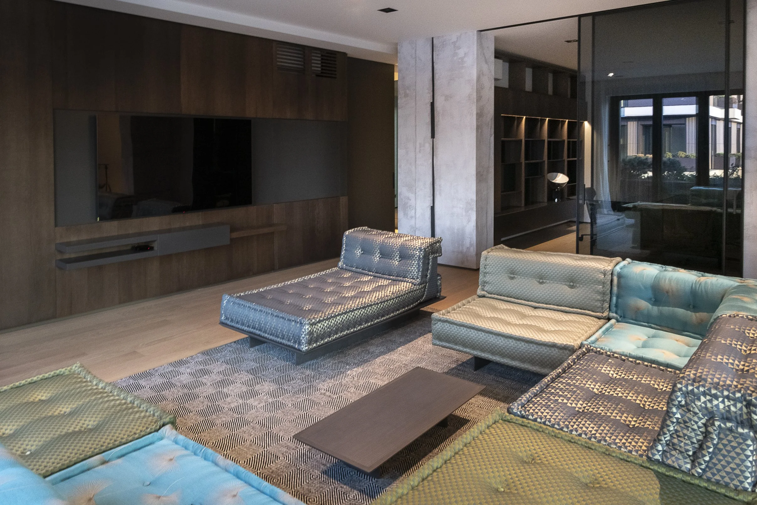 A modern living room with multiple green and blue cushioned mattresses, a wood-paneled wall with a mounted flat-screen TV, a rug with geometric patterns, and a large window with a view of neighboring buildings and greenery.